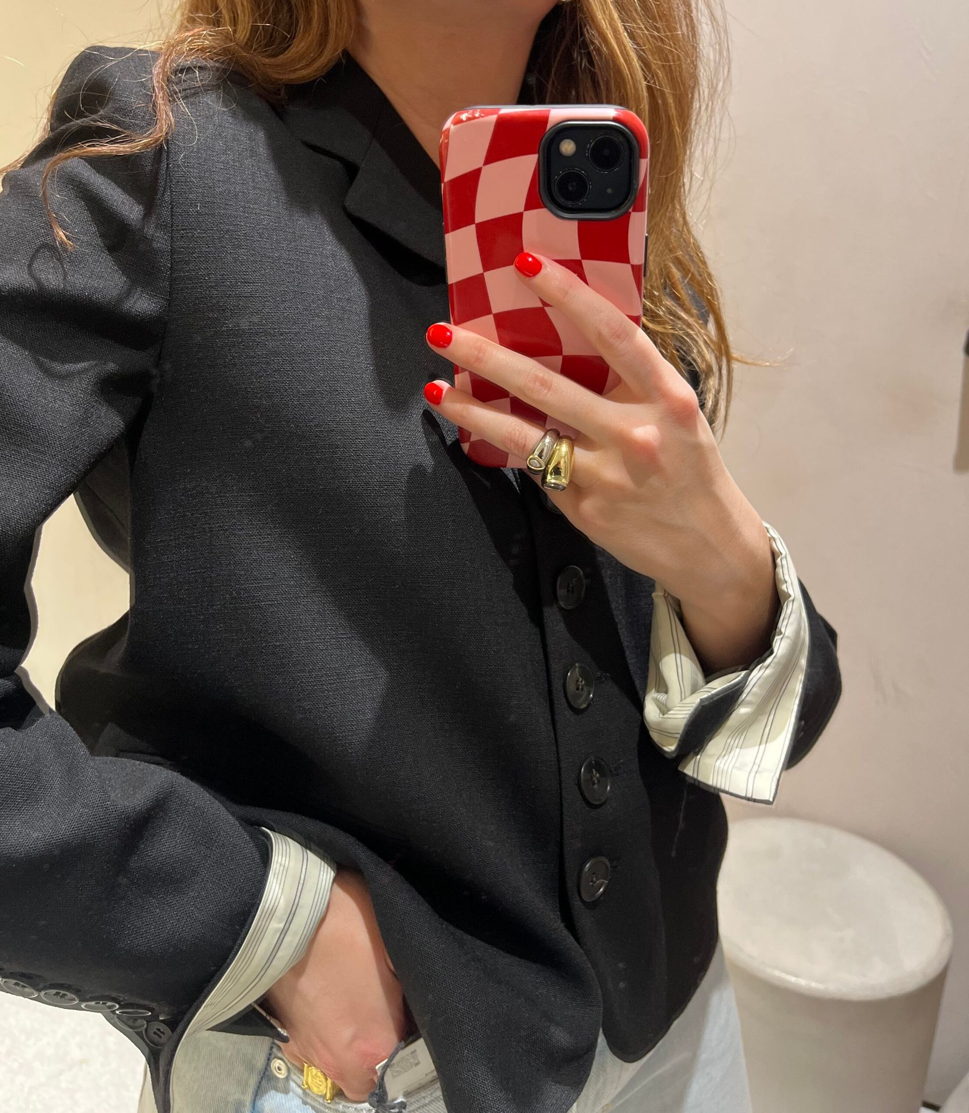 A person with red nail varnish takes a mirror selfie, wearing a black blazer over a striped shirt—both pieces looking new in at Zara. Their phone has a red and white chequered case, and gold rings are visible on their fingers.