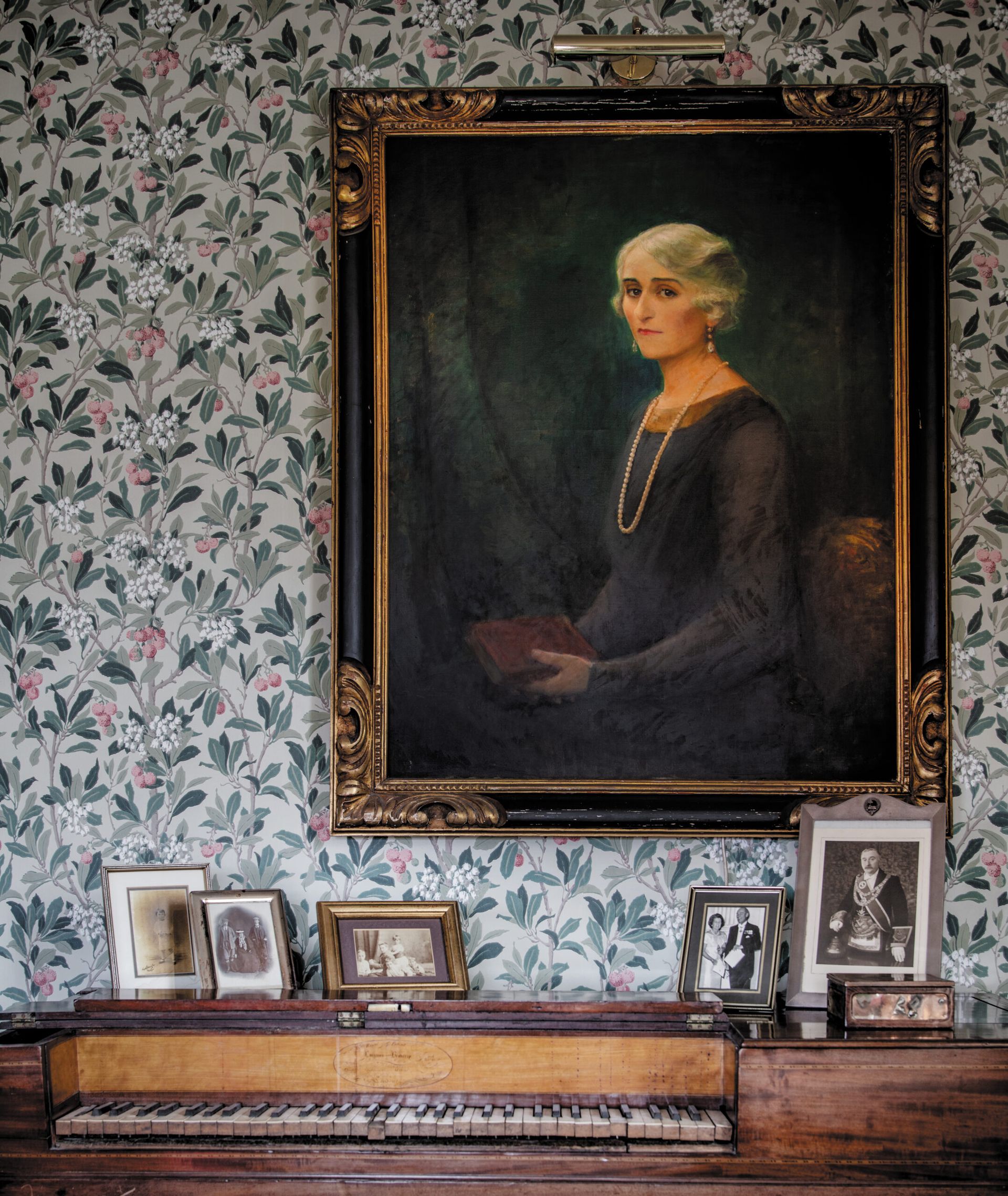 A large portrait of an elderly woman with white hair, wearing a dark dress and pearl necklace, hangs above a vintage piano at Kinloch Lodge. The wall is covered in floral wallpaper, and framed black-and-white photos sit on the closed piano lid.