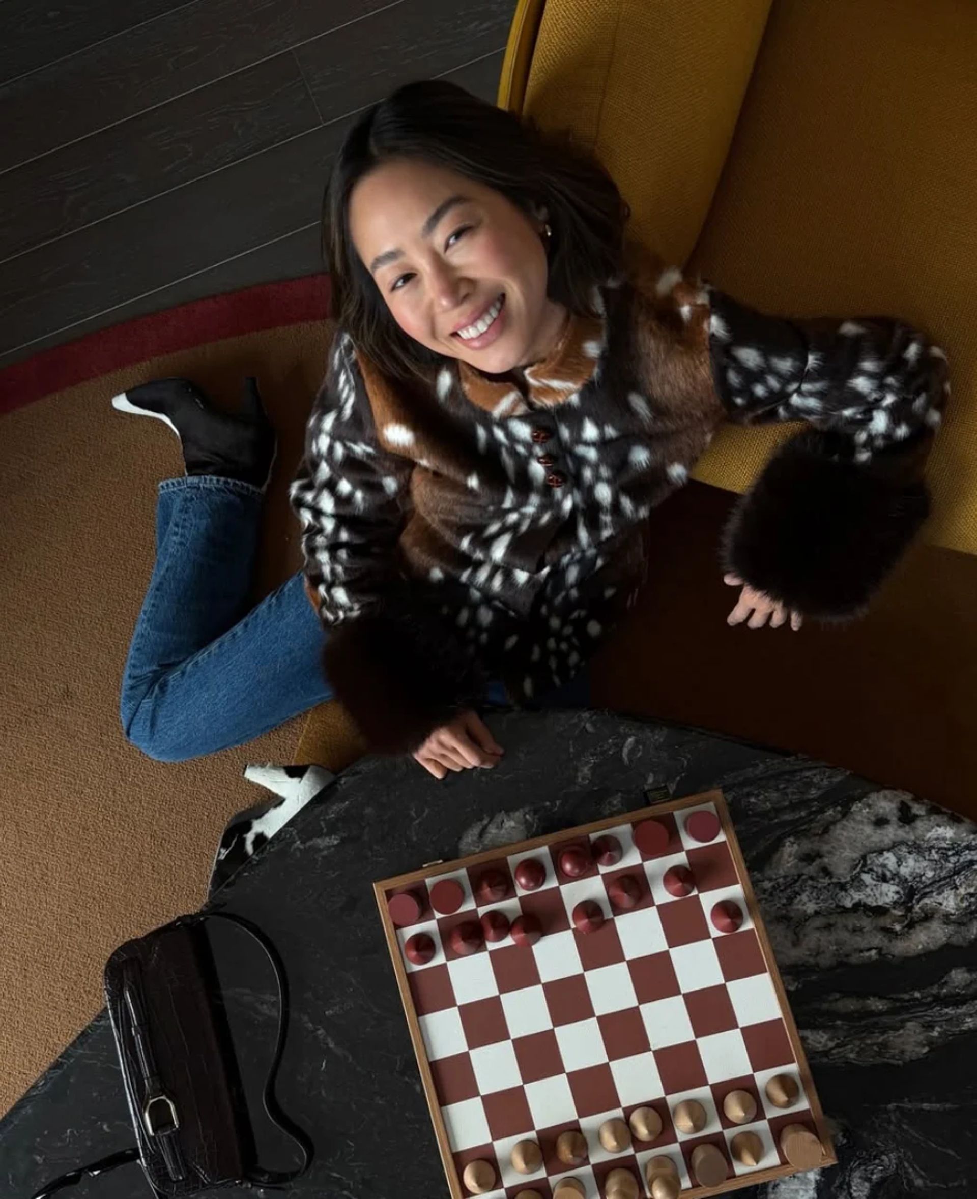 Woman smiling, sitting on the floor beside a black handbag from one of her favourite small fashion brands and a chessboard with wooden pieces, wearing jeans, a patterned fur jacket, and black shoes, with a yellow chair and dark floor visible.