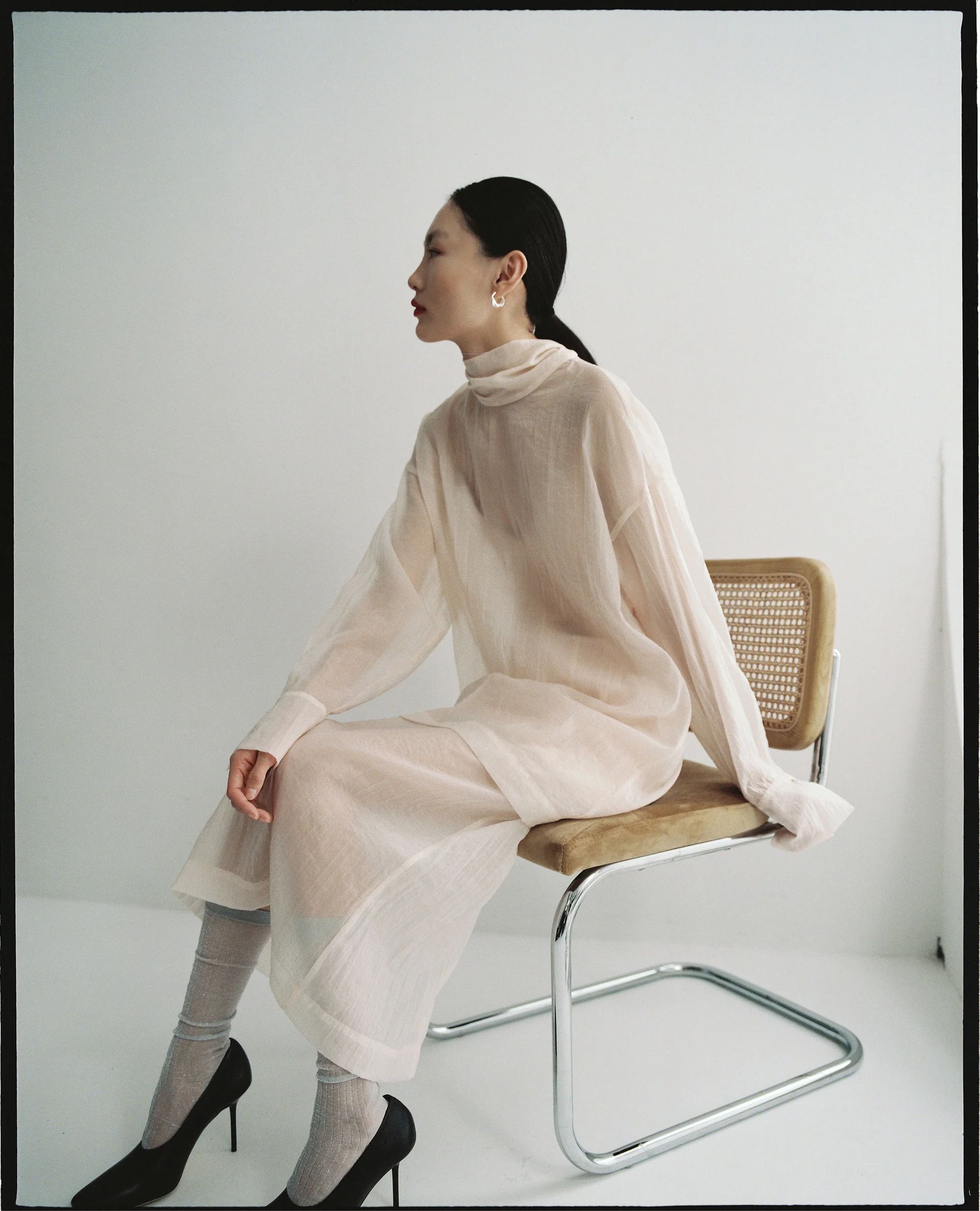 A woman in a sheer, long white dress and black heels from small fashion brands sits sideways on a modern chair, looking left. The background is plain and minimalistic.