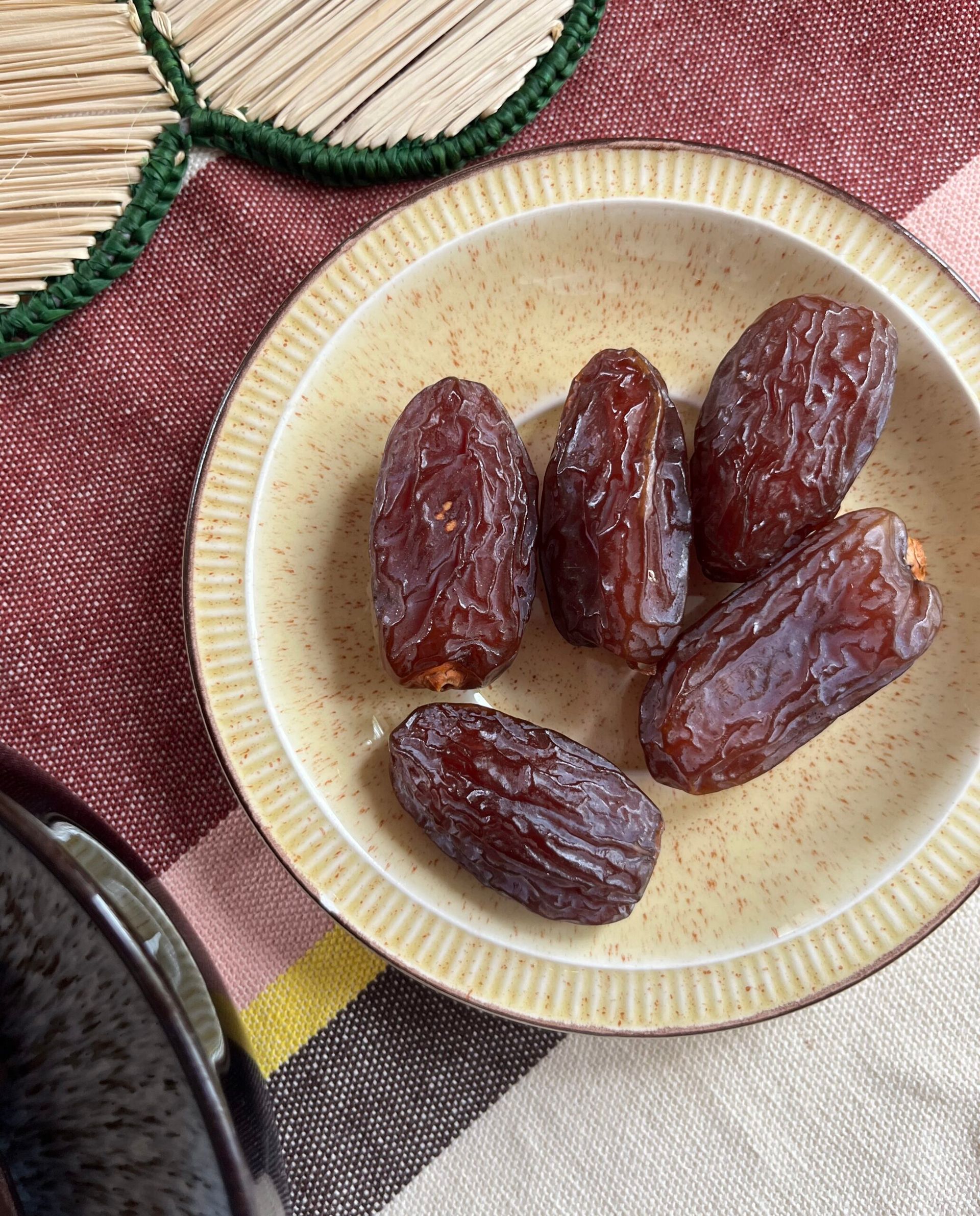 A small beige plate holds five plump, glossy peanut butter dates on a table with a red-brown cloth and a woven mat nearby.
