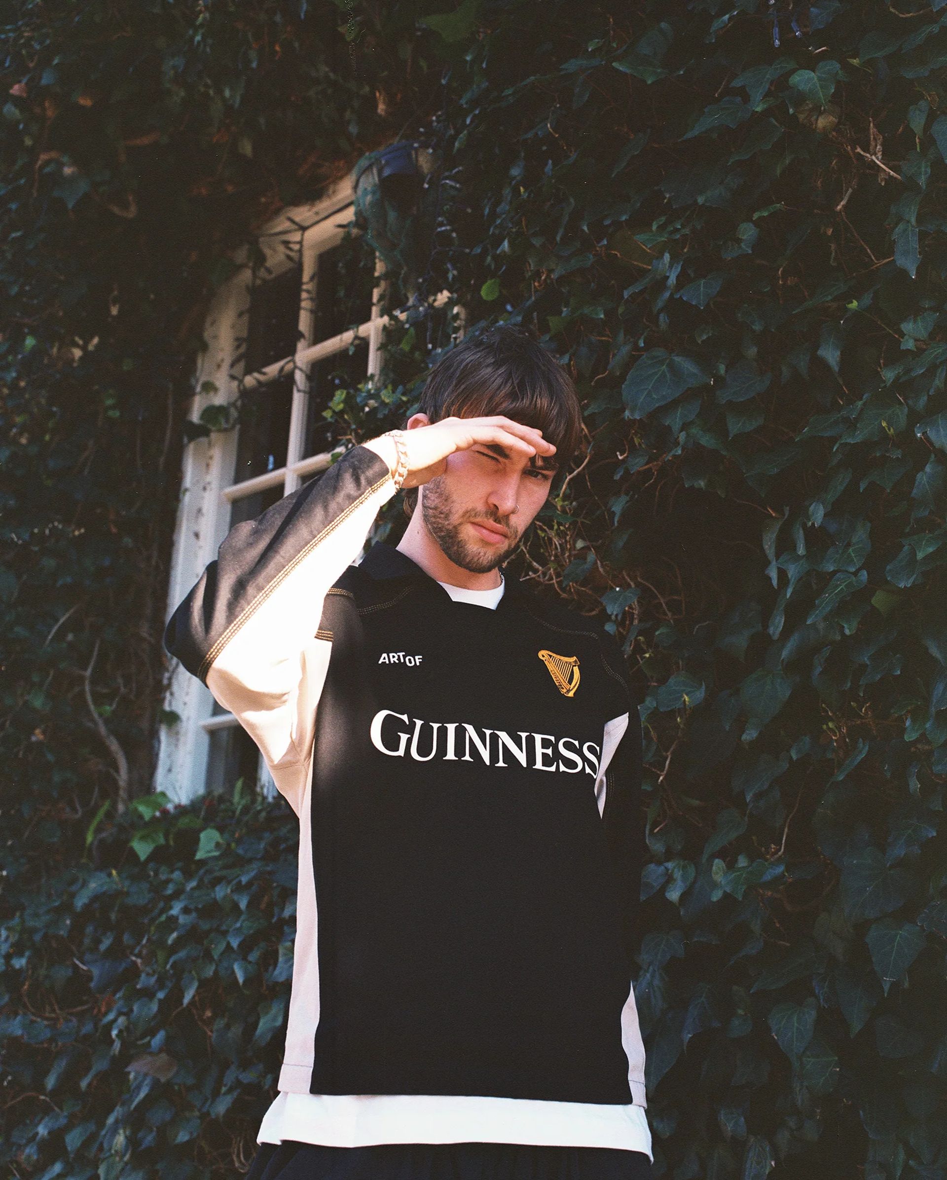 A man in a black Guinness shirt, channelling football core style, stands outdoors before an ivy-covered window, raising his hand to shield his eyes from the sunlight.