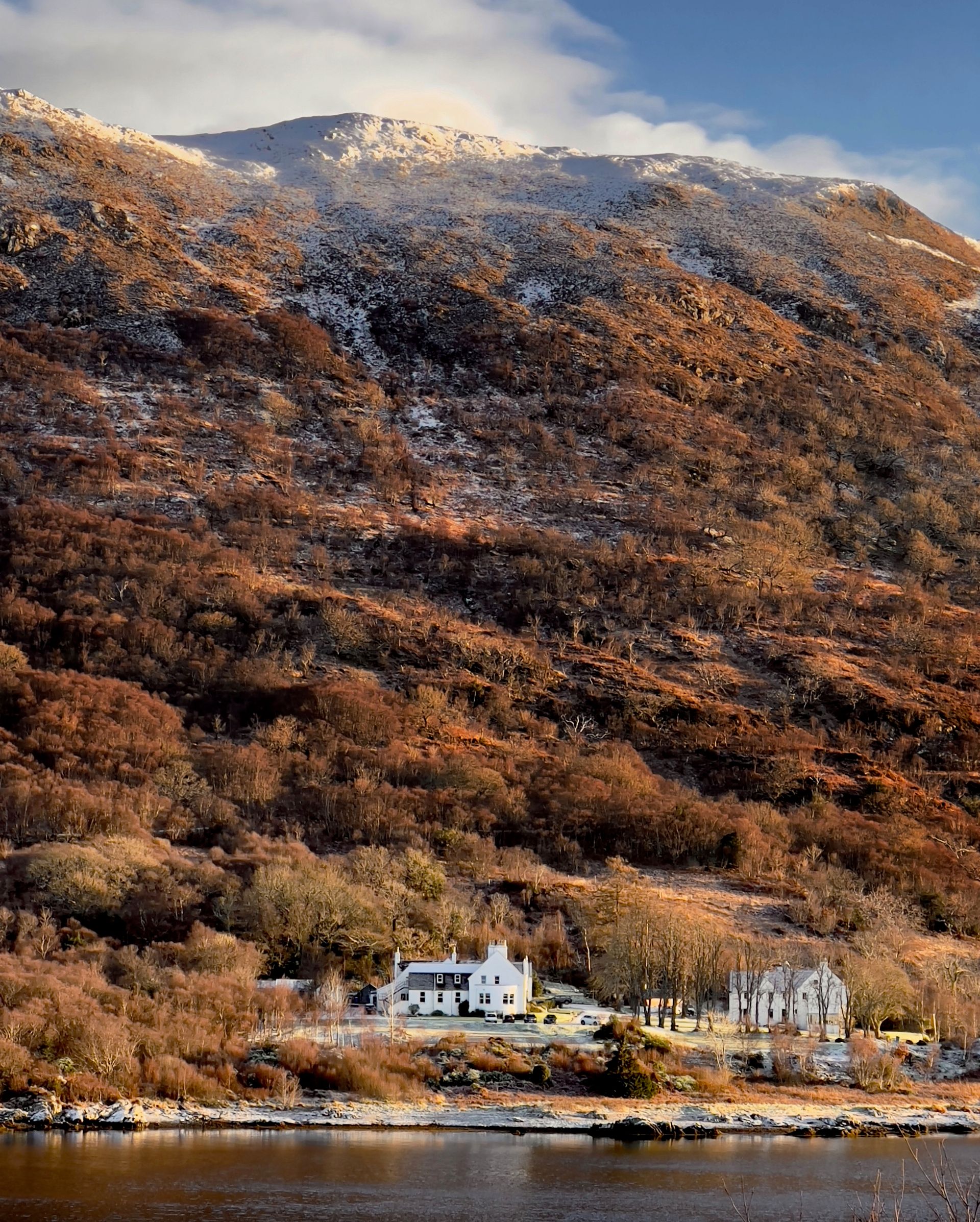 A white house, reminiscent of Kinloch Lodge, sits beside a lake at the base of a large, brown, tree-covered hill dusted with snow under a partly cloudy sky. The tranquil scene is bathed in warm sunlight.