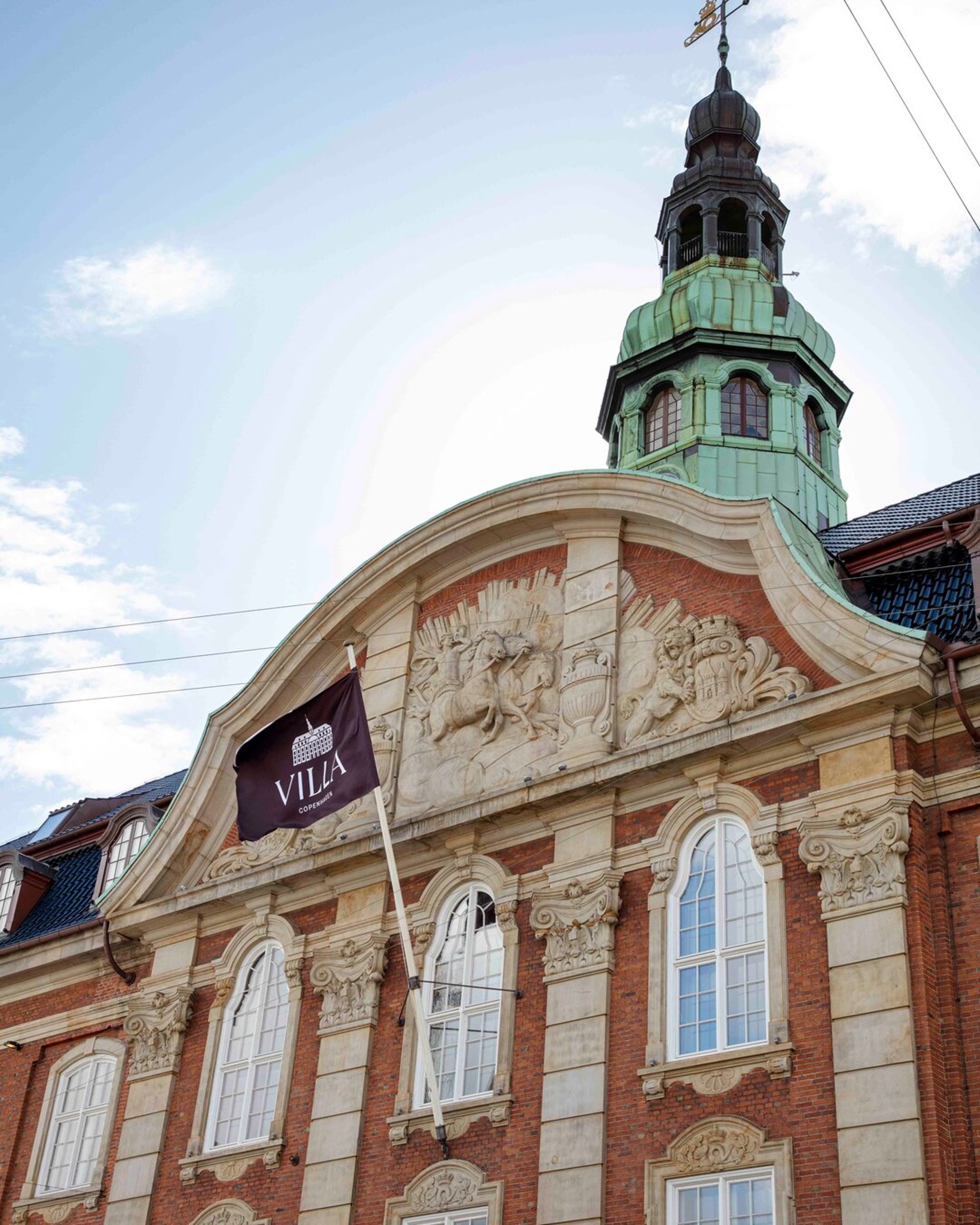 Historic brick building with ornate stone carvings above arched windows, a green copper dome, and a flag reading Villa Copenhagen hanging from the facade. Perfect for your 2026 travel plans. The sky is partly cloudy in the background.