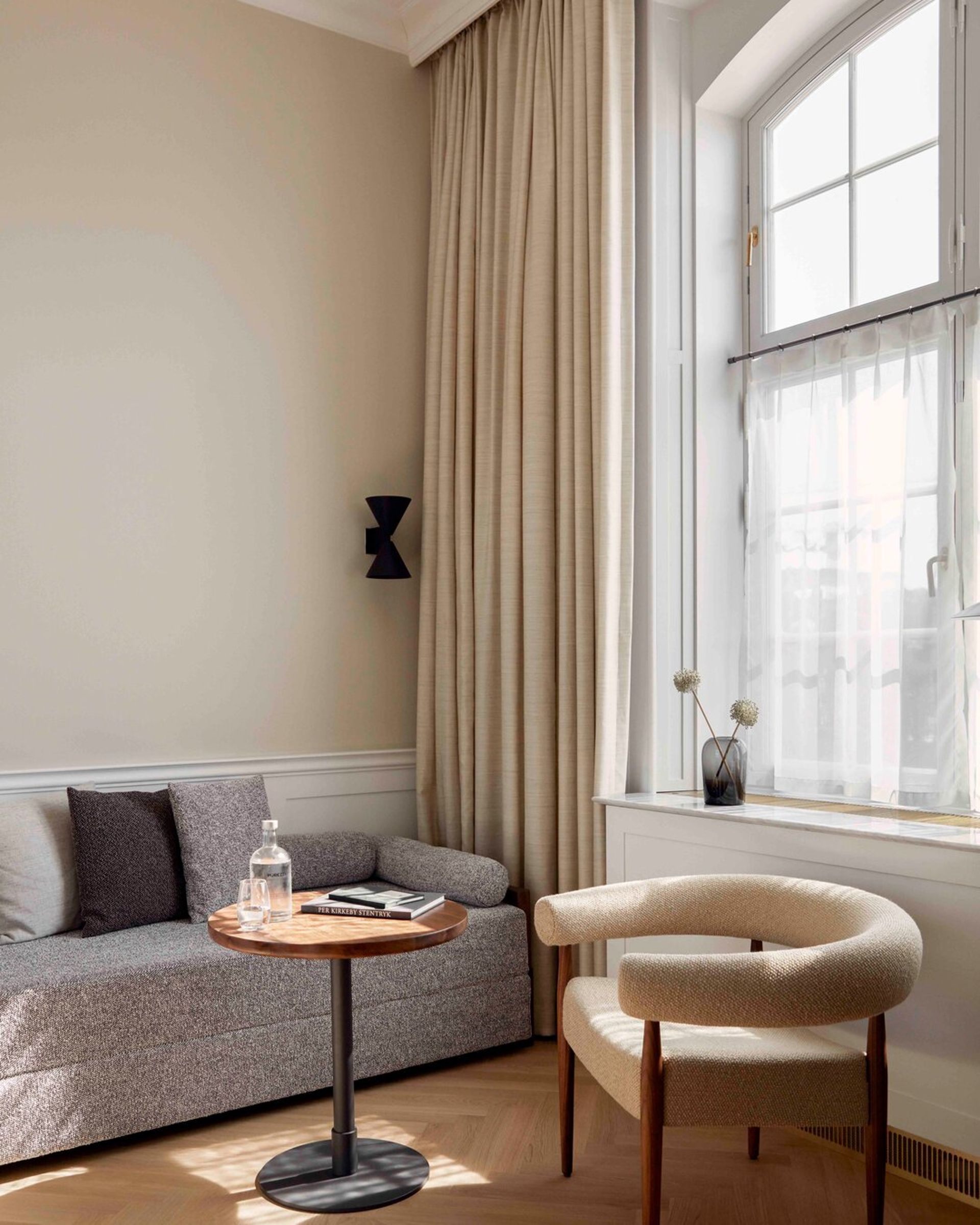 A cosy, minimalist living room inspired by Copenhagen style features a grey sofa, round wooden side table, modern cream armchair, beige curtains, and a large window. A glass bottle and book sit on the table, evoking the charm of Villa Copenhagen.