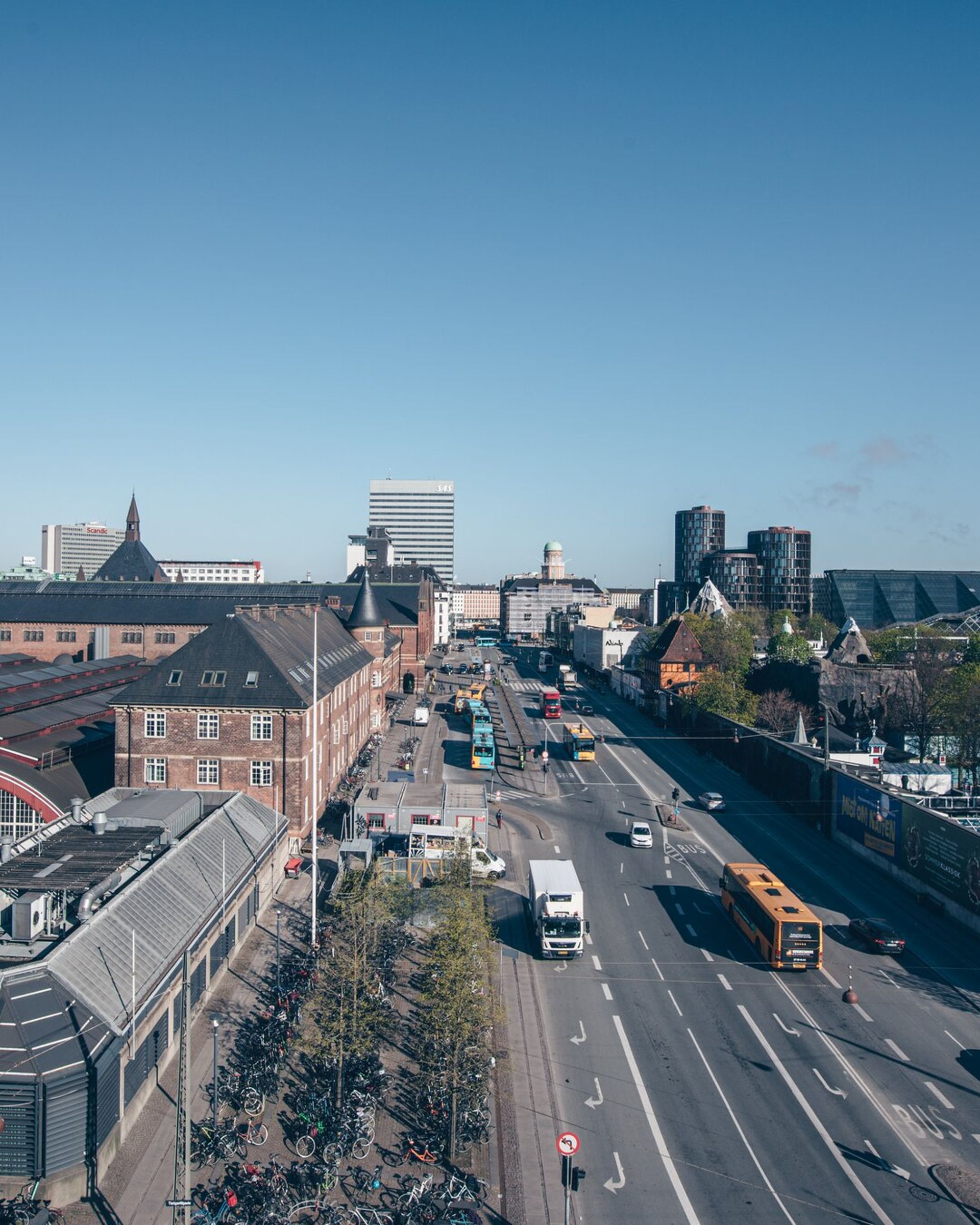 A wide city street in Copenhagen with cars, buses, and bicycles runs between buildings under a clear blue sky. Modern high-rises rise in the background, while bikes are parked along the pavement—perfect for your 2026 travel adventures near Villa Copenhagen.