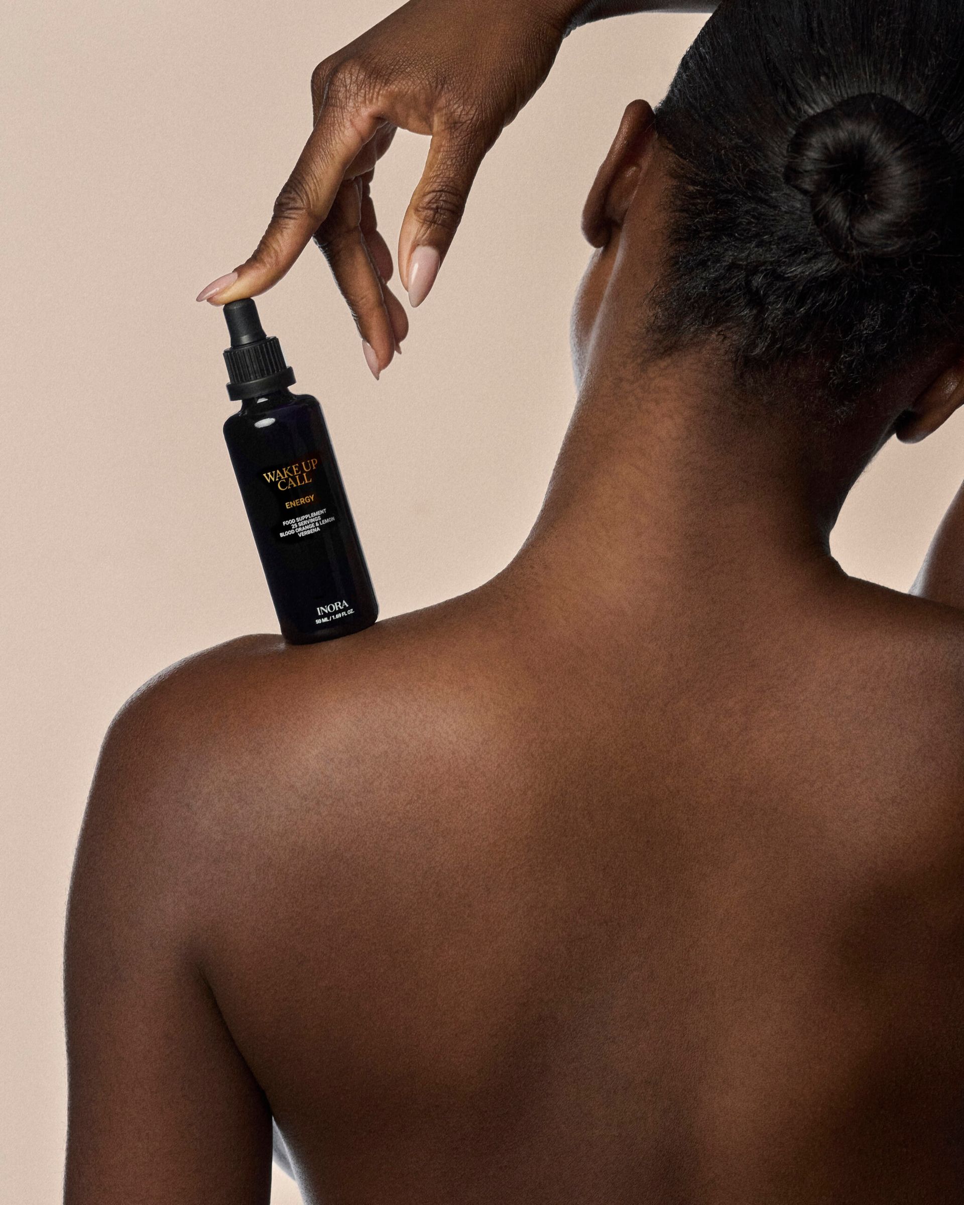 A person with dark skin and hair in a bun is shown from behind, channelling London Cool Girls style as they hold a black bottle of Wealt Lip Elixir Serum on their shoulder against a beige background. Have It All with this chic beauty essential.