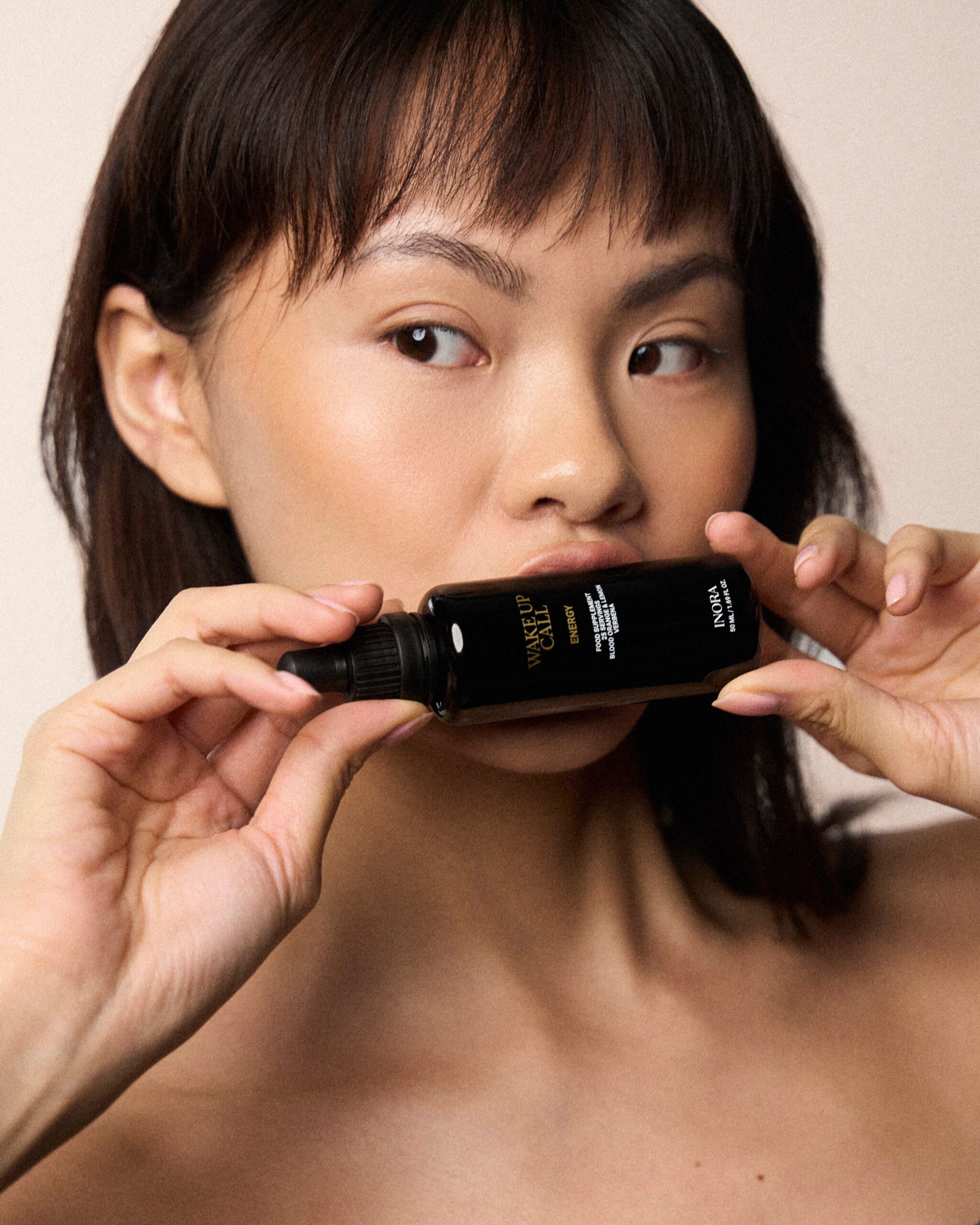 A woman with smooth skin holds a black bottle of skincare serum close to her lips, gazing to the side—a true Have It All moment. The neutral background highlights her glowing complexion and the product, channelling effortless London Cool Girls vibes.