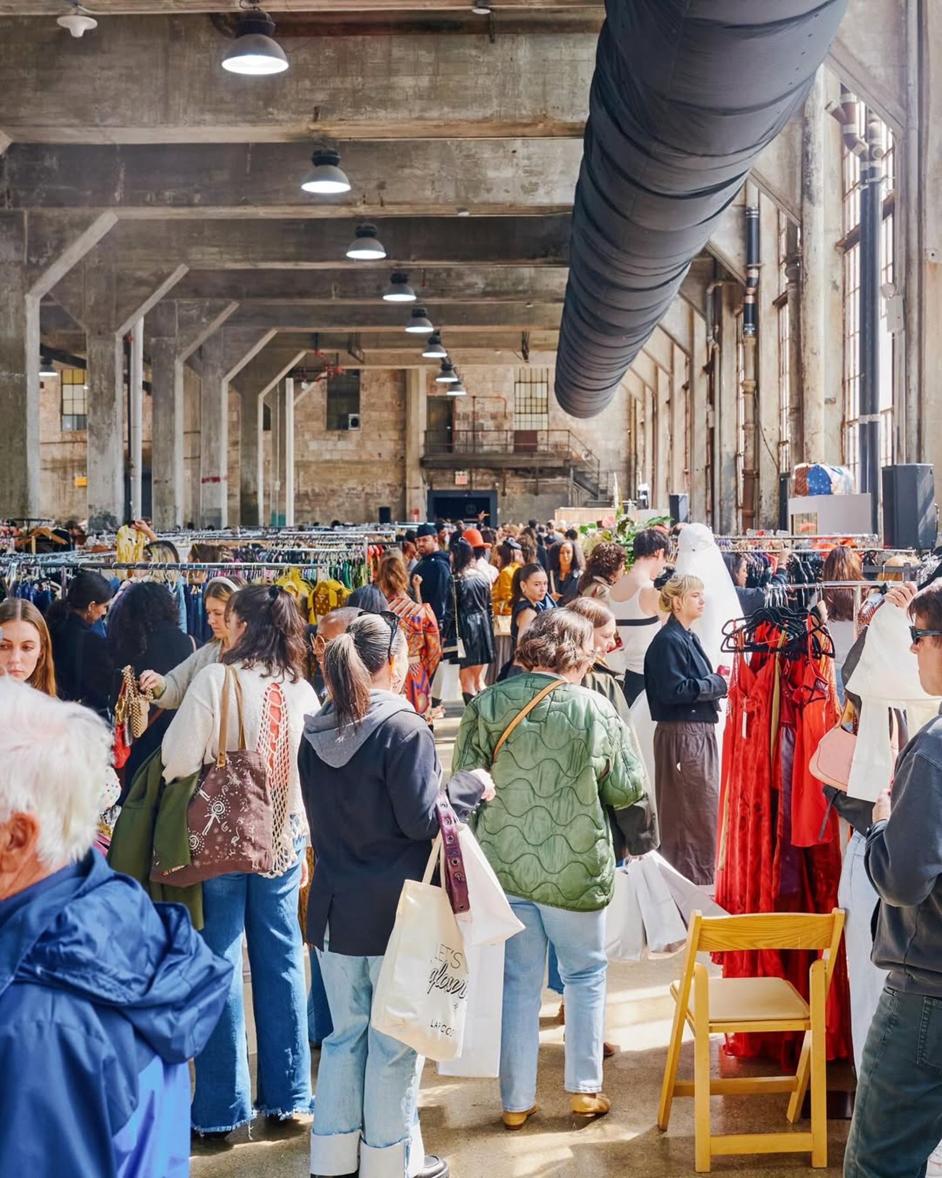 A busy indoor market filled with people browsing rails of clothing in a large, industrial-style building. Sunlight streams through tall windows, making it a top spot for weekend activities and adding to the lively, bustling atmosphere.