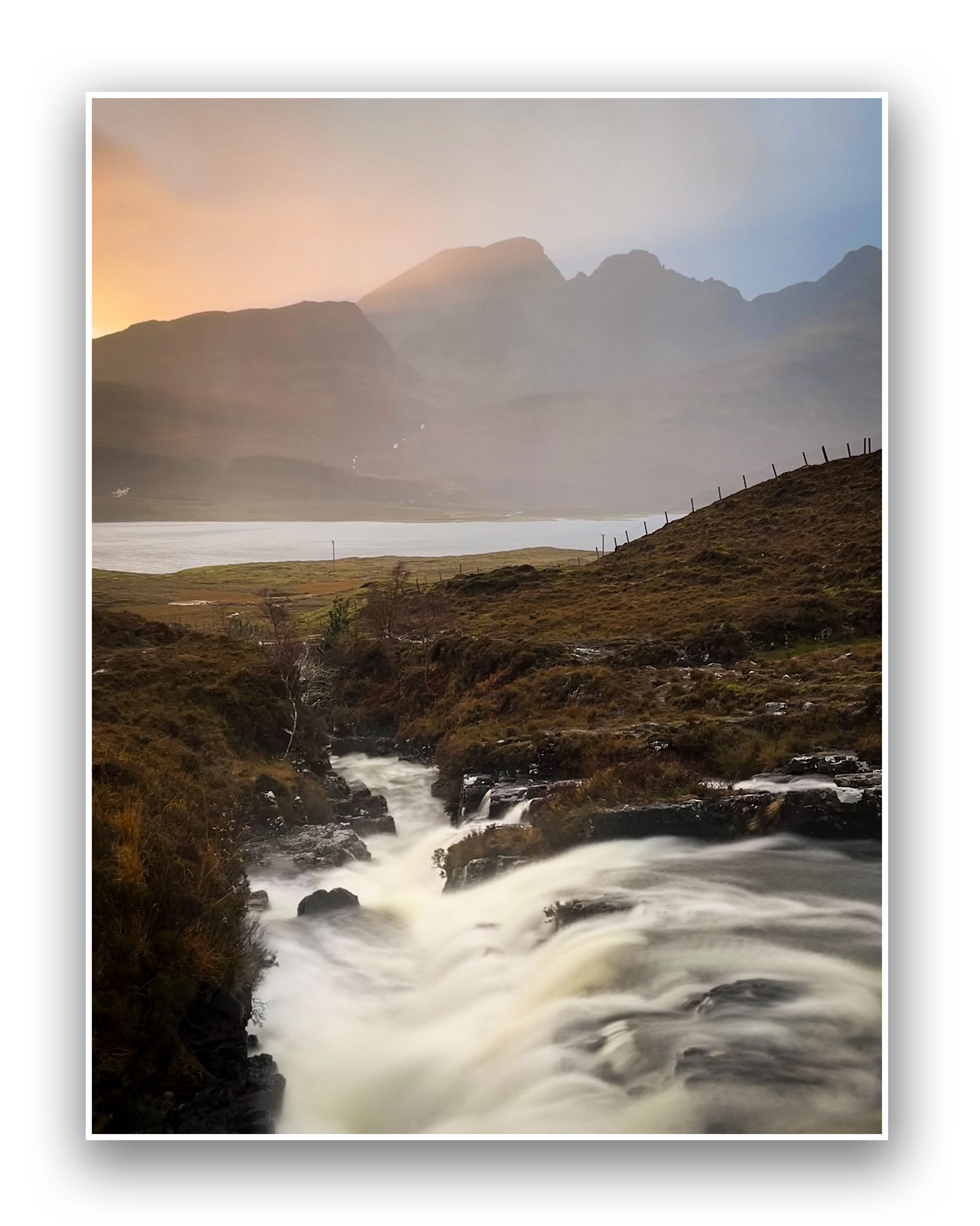 A gentle stream flows over rocks in the foreground, with misty mountains and a calm lake near Kinloch Lodge in the background under a softly coloured sunset sky. A grassy hillside and a rustic fence line the right side.