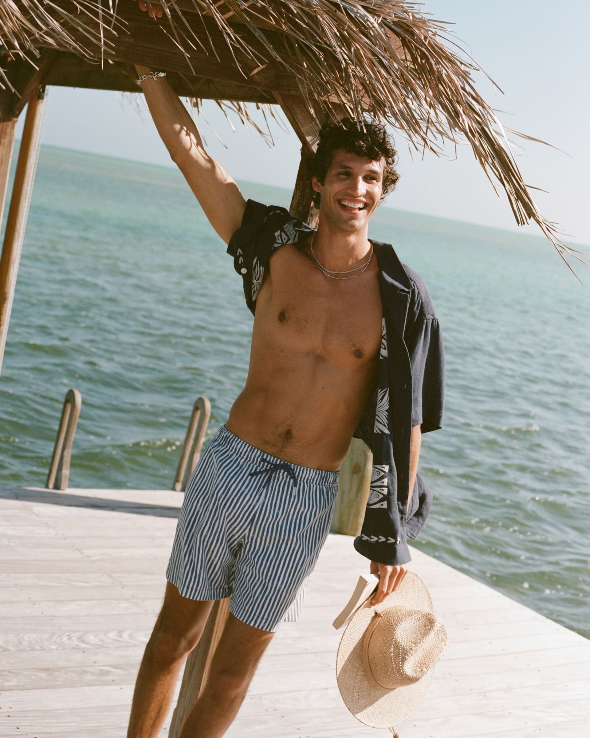 A smiling man in swimming trunks and an open shirt stands on a wooden jetty by the sea, showcasing men's holiday fashion as he holds a straw hat and leans against a thatched roof. Bright water and sky complete the scene.