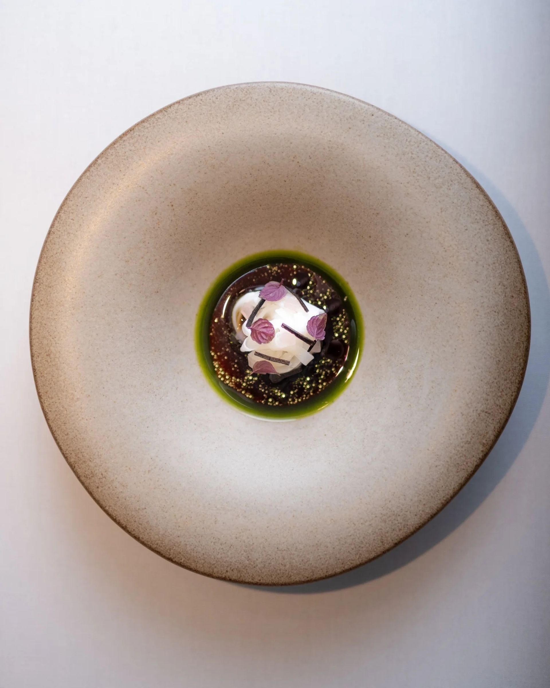 A round beige bowl with a green sauce at the centre, topped with a white sphere garnished with purple flower petals and microgreens—an elegant dish you might discover in an expertly curated Edinburgh travel guide featuring insider tips for the city’s renowned culinary scene.