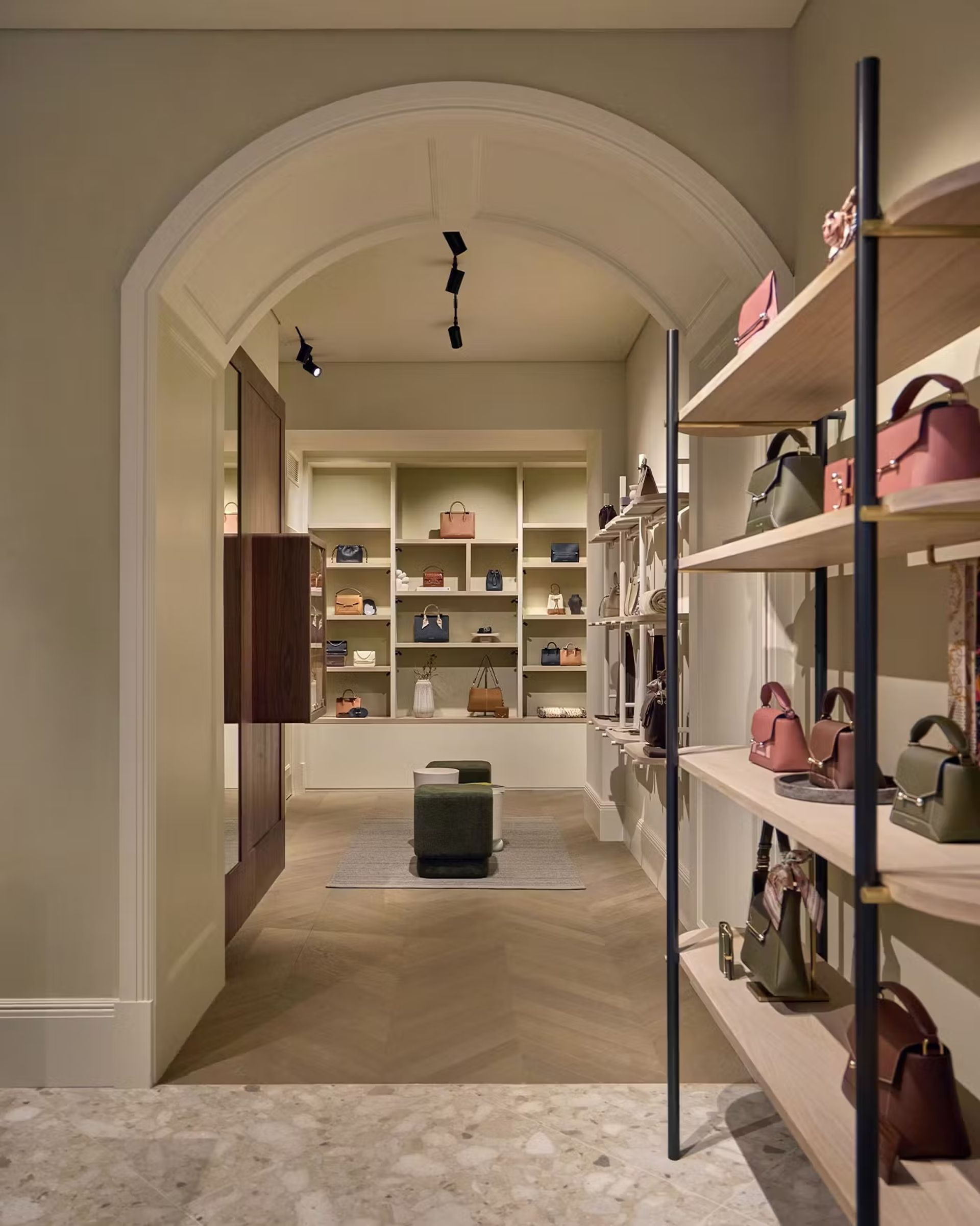 A stylish boutique in Edinburgh displays various handbags on shelves and in wall cubbyholes, with a green stool in the centre beneath an arched doorway and soft lighting—an inviting spot for any fashion-loving traveller seeking insider’s guide-worthy finds.