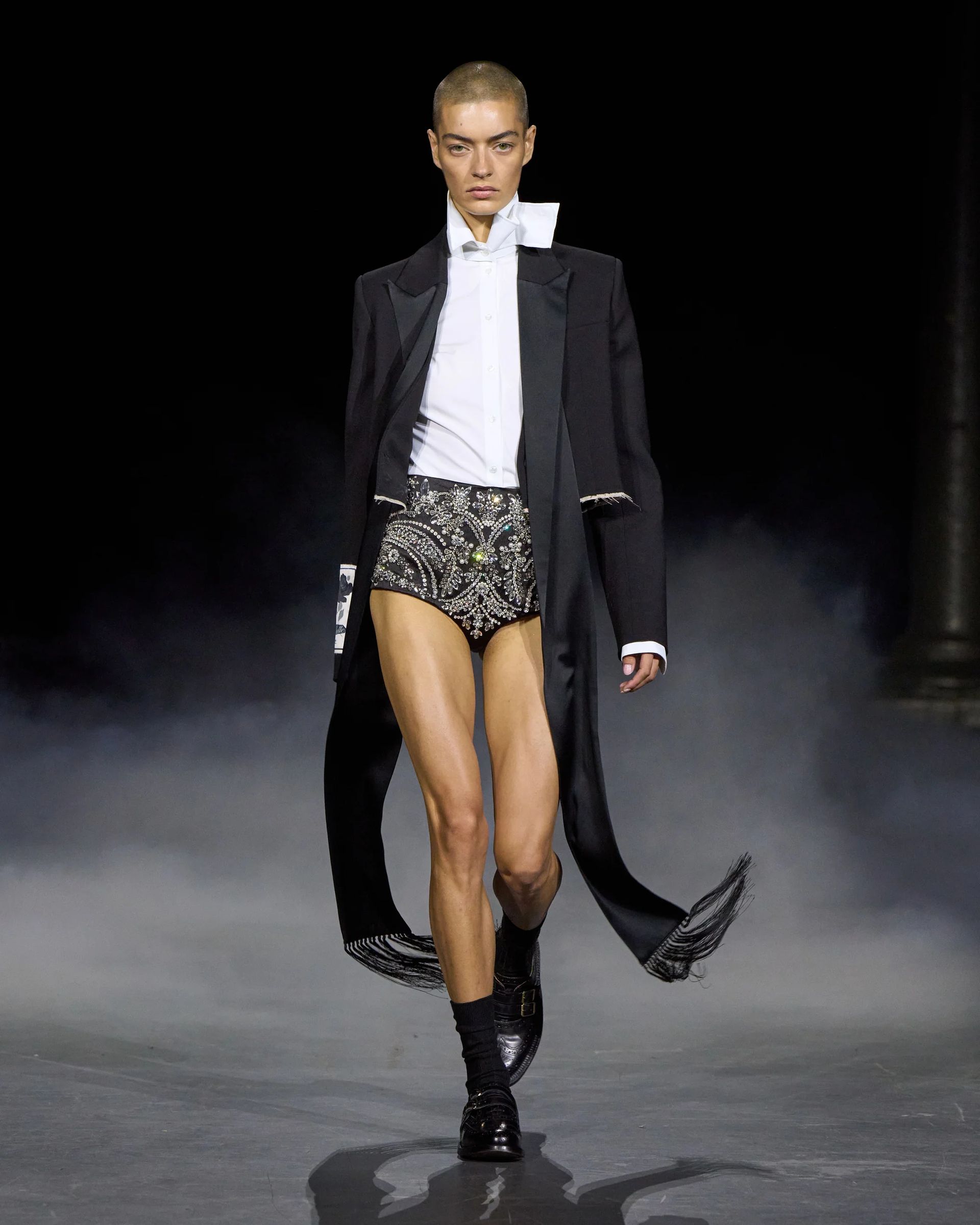A model with a shaven head walks a foggy catwalk at London Fashion Week, wearing a long black coat, white dress shirt, high-collared black shorts adorned with rhinestones, black socks, and black shoes.