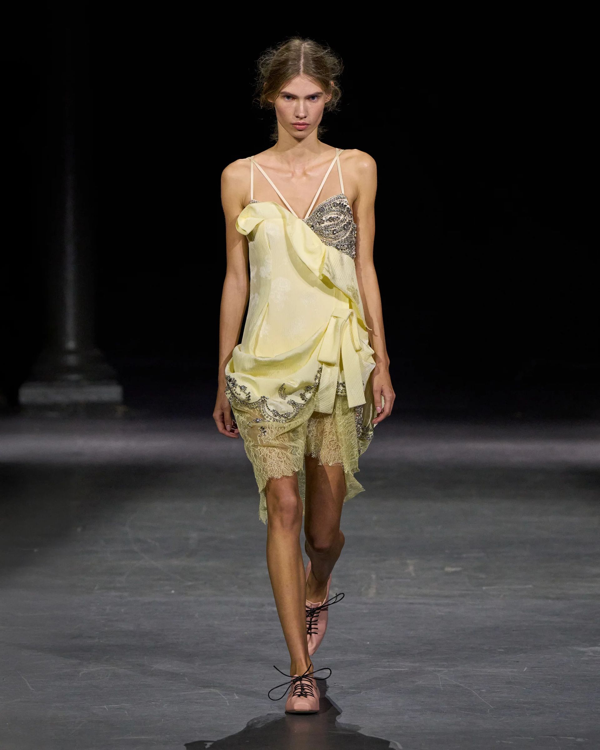 A model walks a catwalk at London Fashion Week wearing a light yellow, asymmetrical dress with lace and frill details, thin straps, and flat pink shoes, against a dark background.