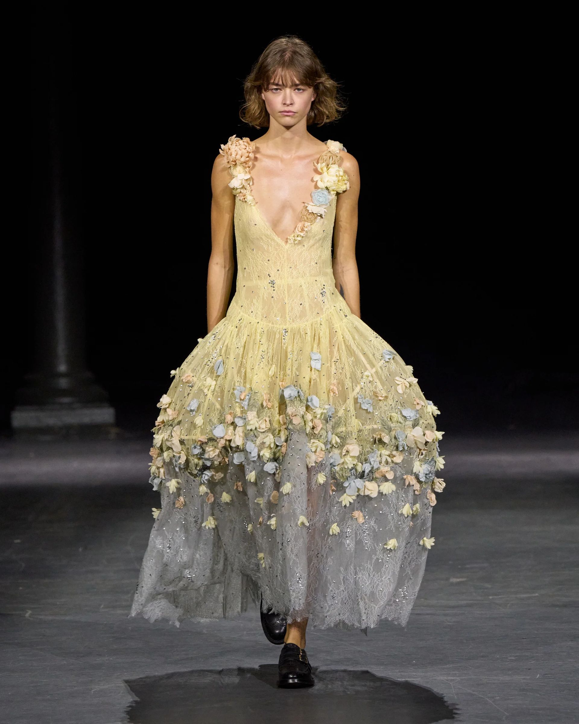 A model walks a catwalk at London Fashion Week wearing a sleeveless, pale yellow dress adorned with 3D floral appliqués, a deep V-neck, and a sheer, flowing skirt, paired with black shoes against a dark background.