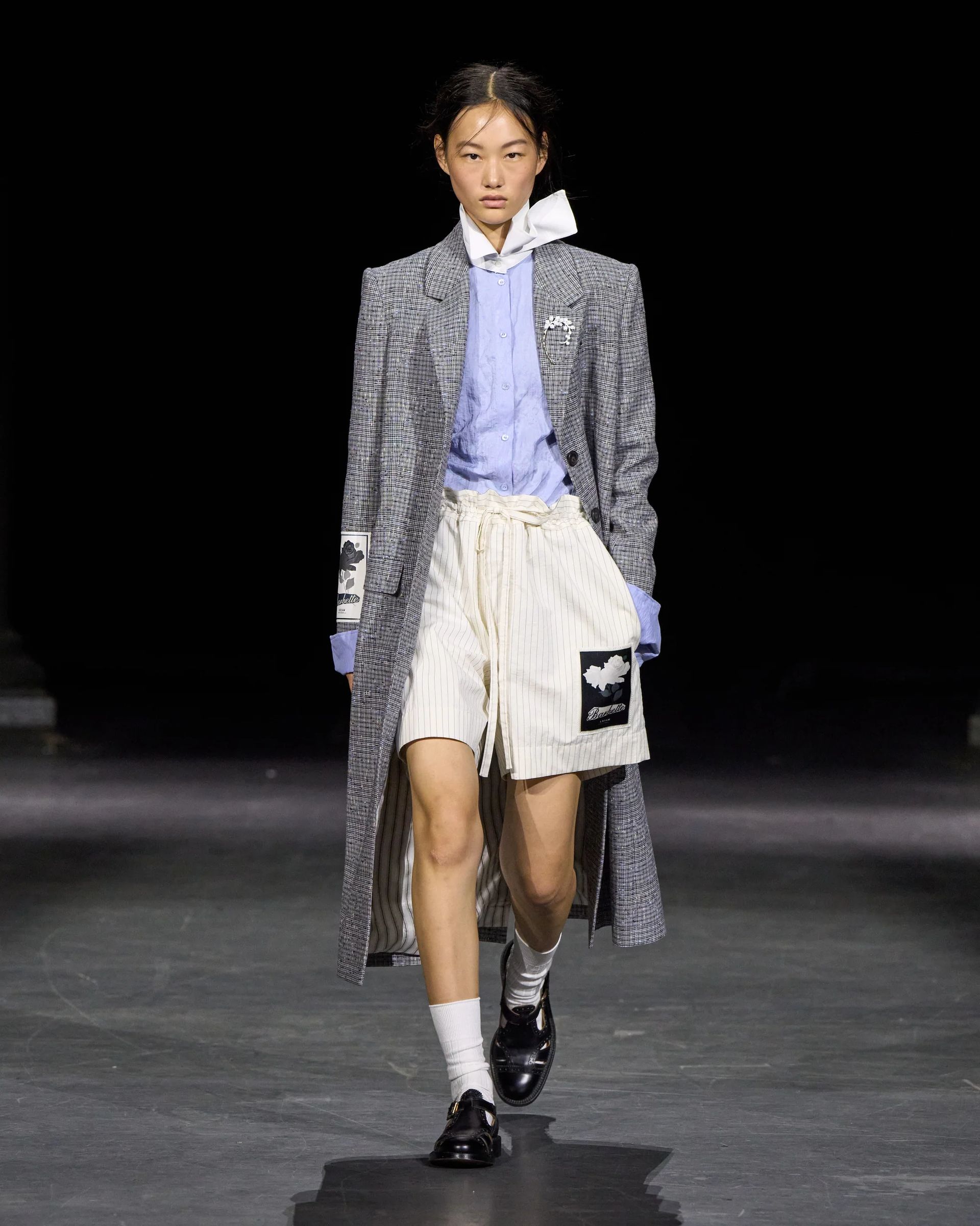 Model walks catwalk at London Fashion Week wearing a checked long coat, blue button-up shirt, white tie, beige drawstring shorts with patches, white socks, and black shoes, against a dark background—a look sure to catch the eye of any fashion editor covering their favourite shows.