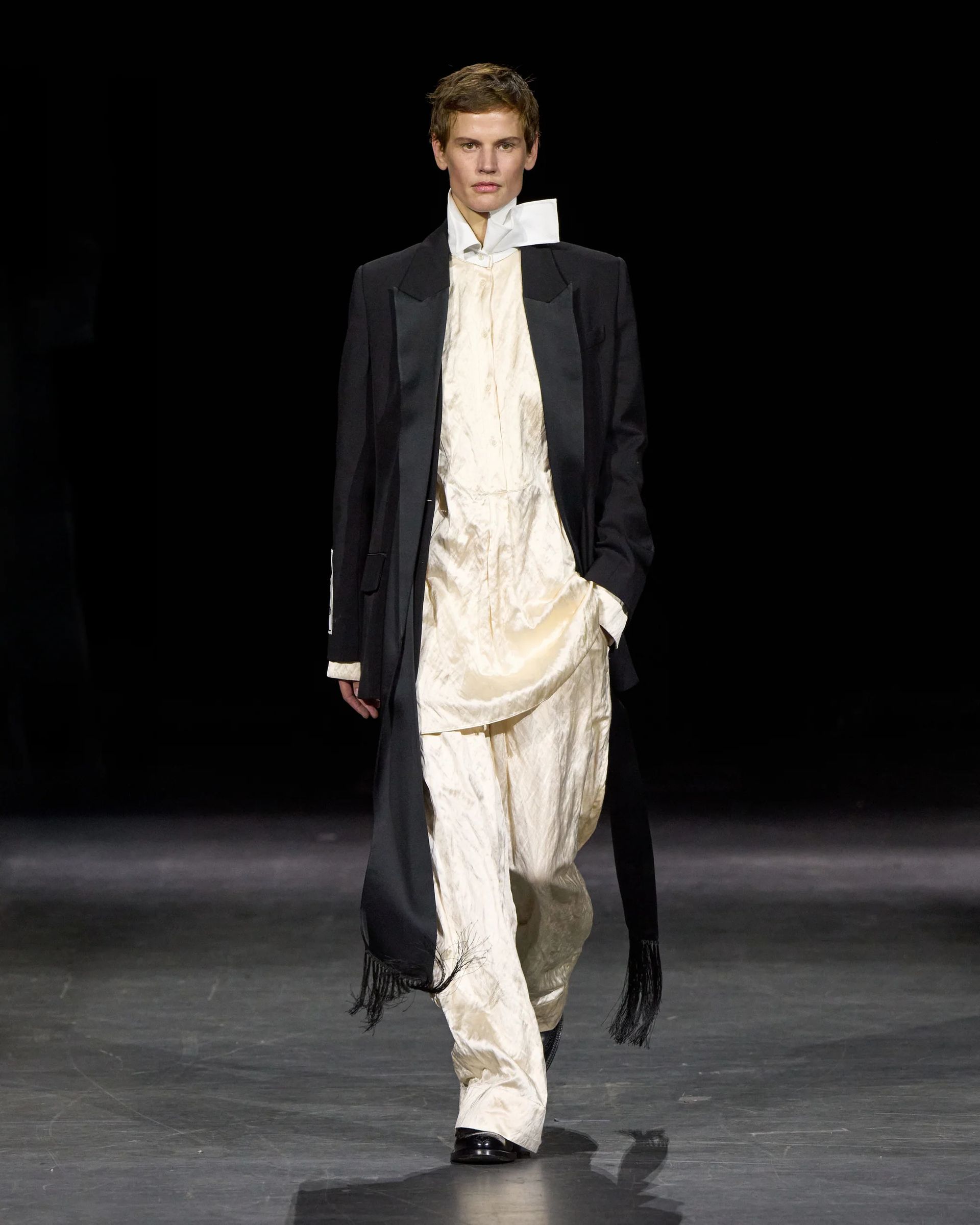 Model walks on a catwalk at London Fashion Week wearing a long black coat over a loose, cream-coloured satin outfit with wide trousers, white boots, and a dramatic tall collar against a dark background—an ensemble that would capture the attention of any discerning fashion editor.