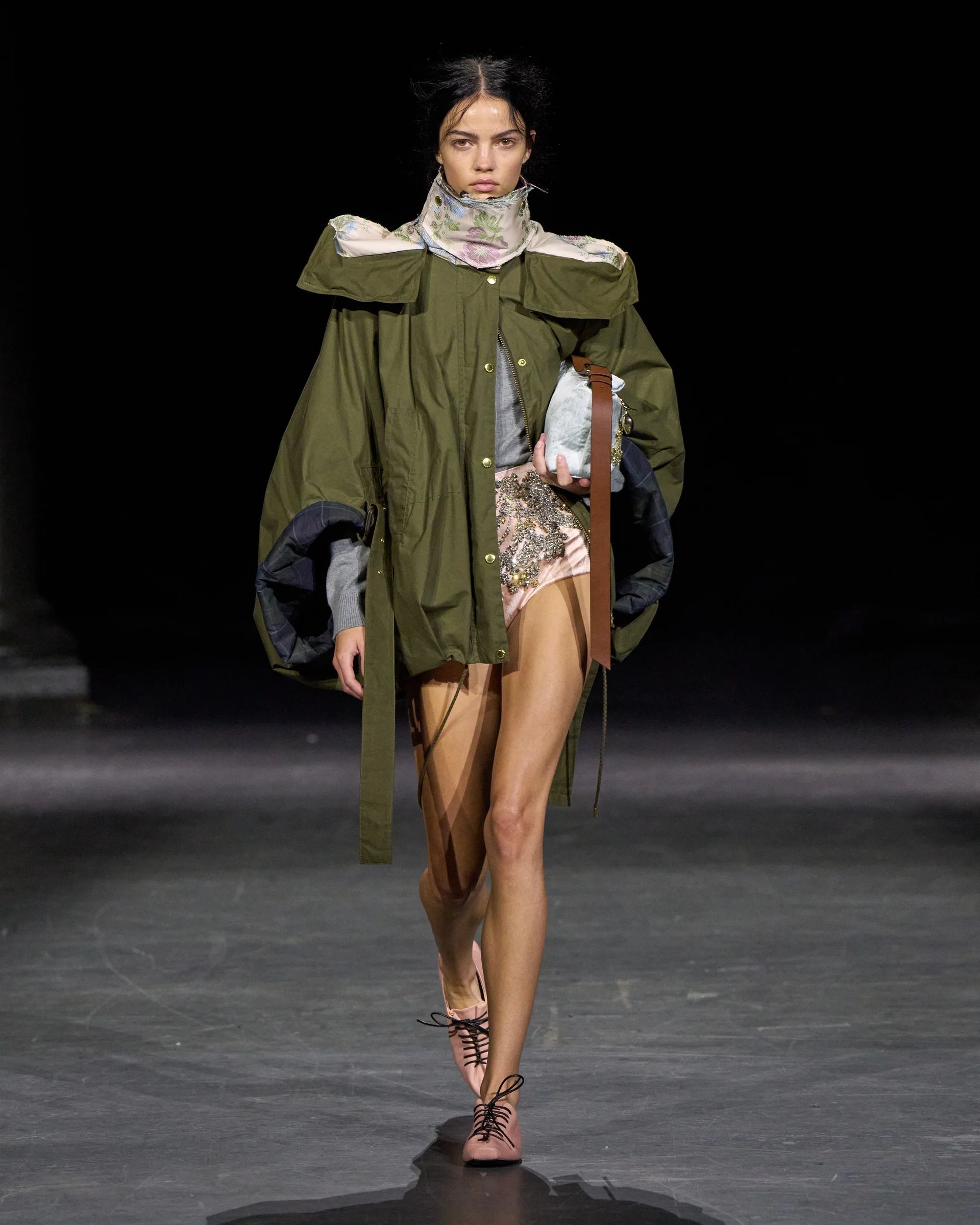 A model walks a catwalk at London Fashion Week wearing an olive green jacket with oversized sleeves, a floral scarf, a silver top, sequin shorts, and light pink lace-up sandals against a dark background.