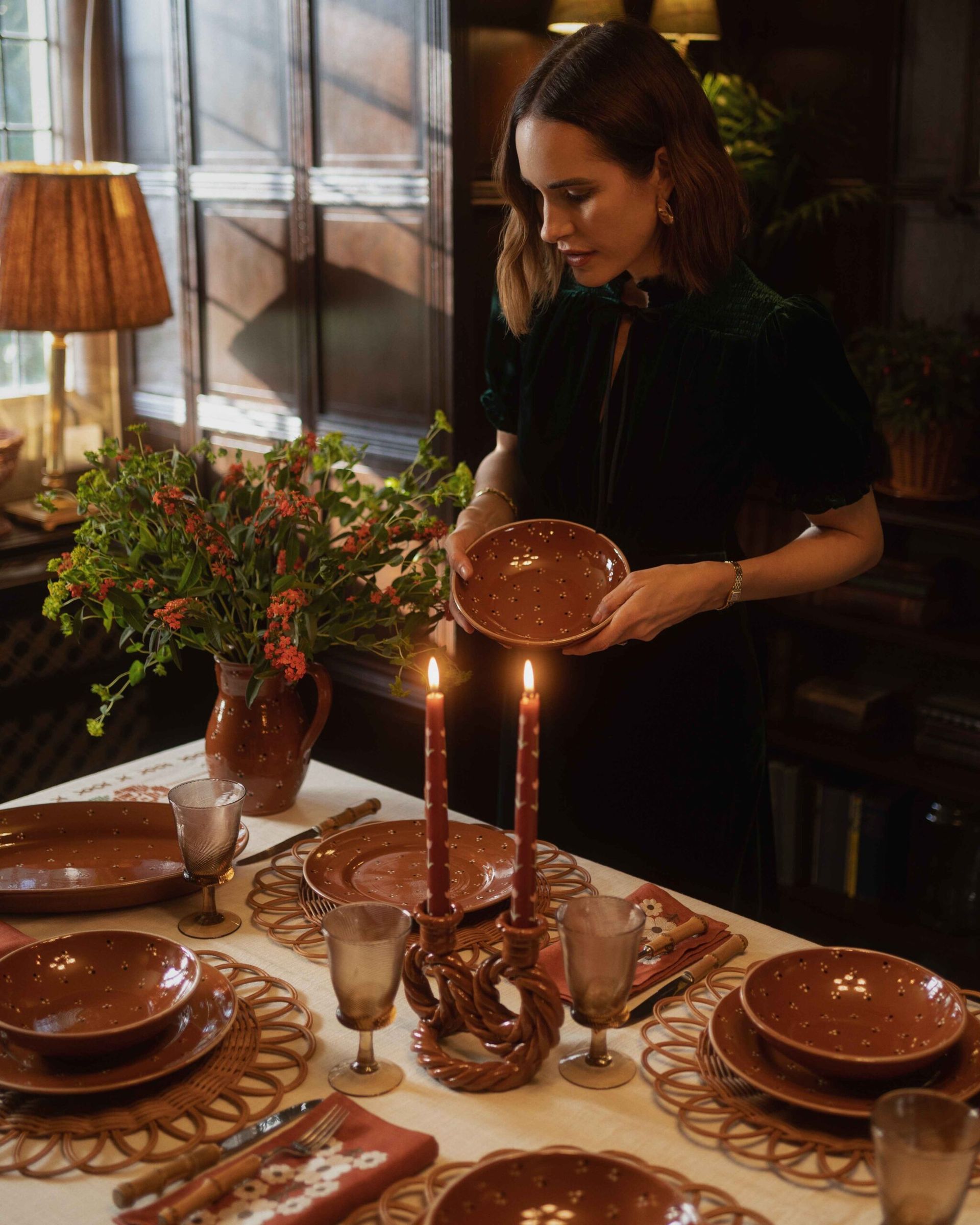A woman in a dark dress sets a speckled brown ceramic plate onto a table arranged for dinner with matching crockery, glasses, and floral-patterned serviettes, offering inspiration for autumn tablescape ideas.