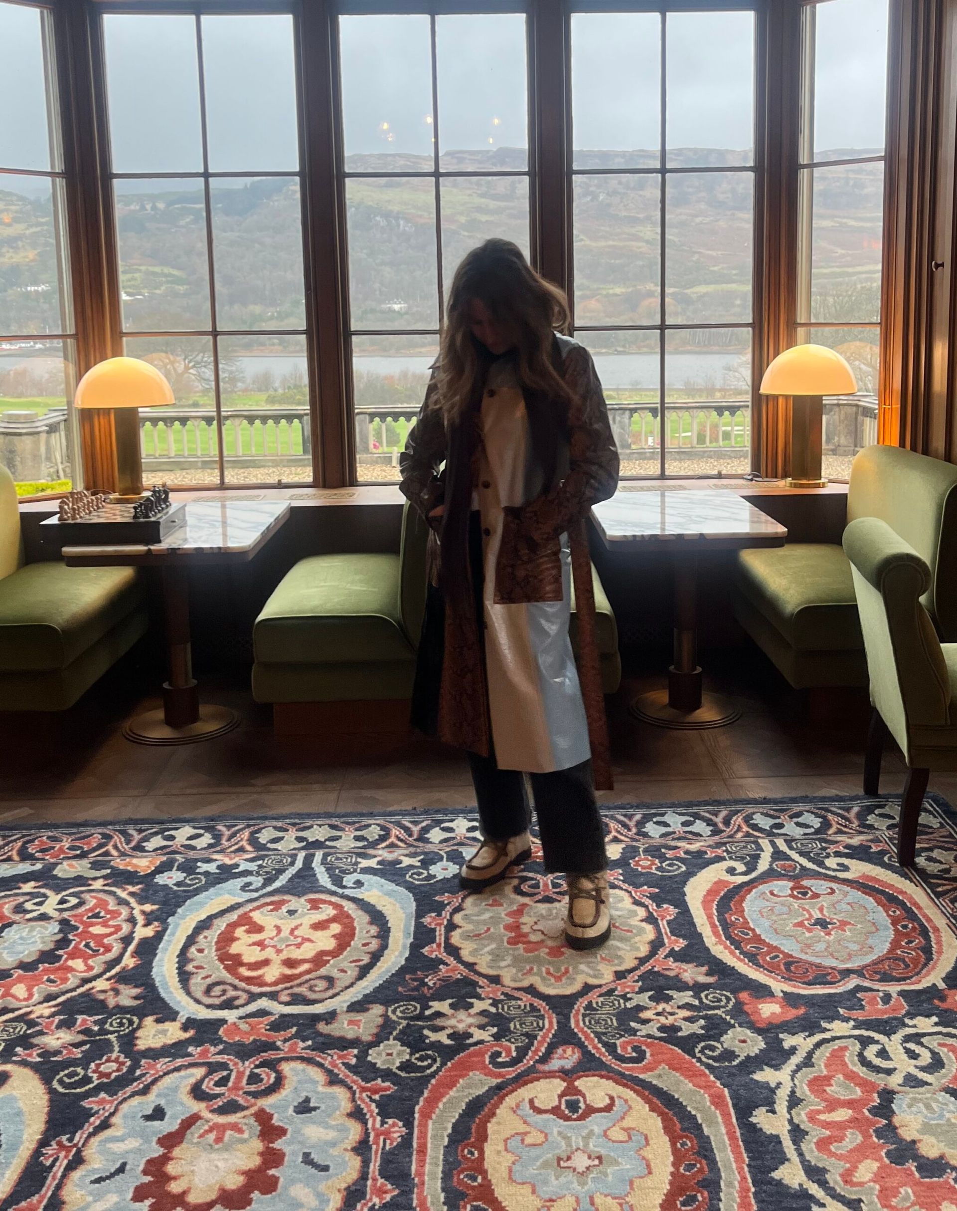 A person in a long coat stands on a patterned rug in a cosy room with green seating, large windows, and scenic hills outside on a cloudy day. Warm lamps sit by the windows, whilst vintage decor and designer dupes add character to the space.
