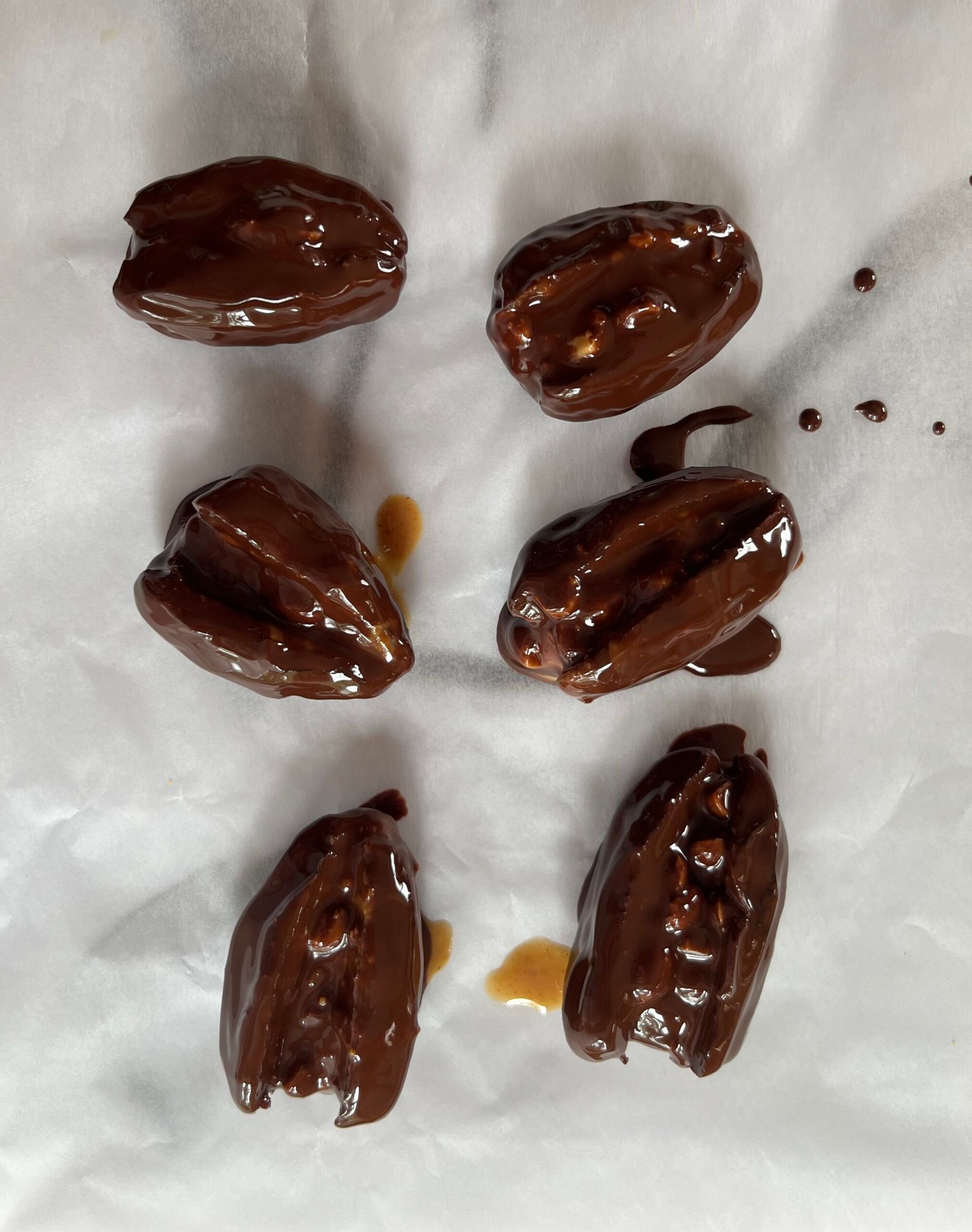 Six peanut butter dates coated in glossy melted chocolate are arranged on white baking parchment, with a few drips of chocolate and caramel-coloured liquid visible nearby.