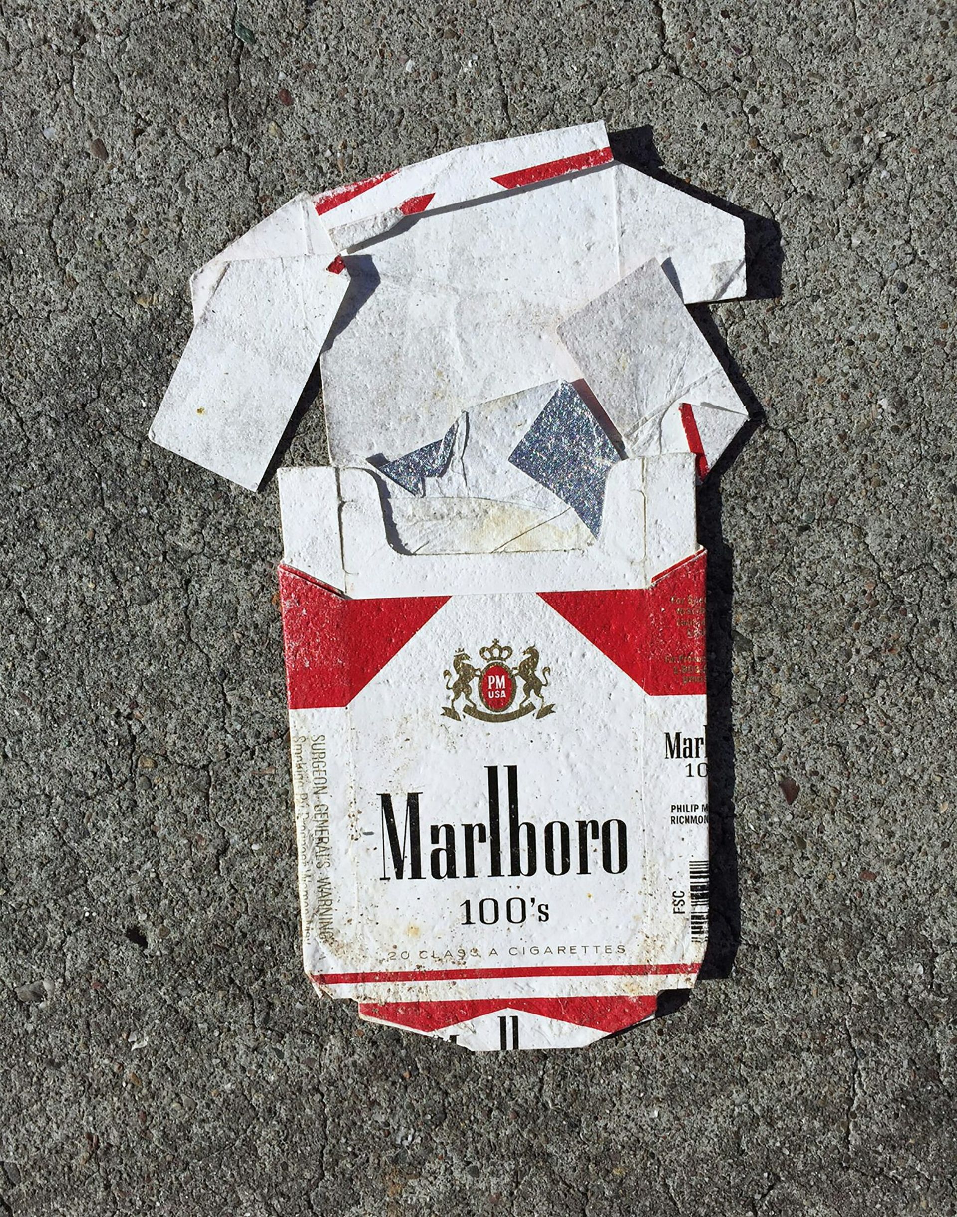 A flattened Marlboro 100’s cigarette box lies on rough grey pavement, its red and white design weathered and creased—a quiet remnant amid rising discussions about smoking rates and the new smoking law.