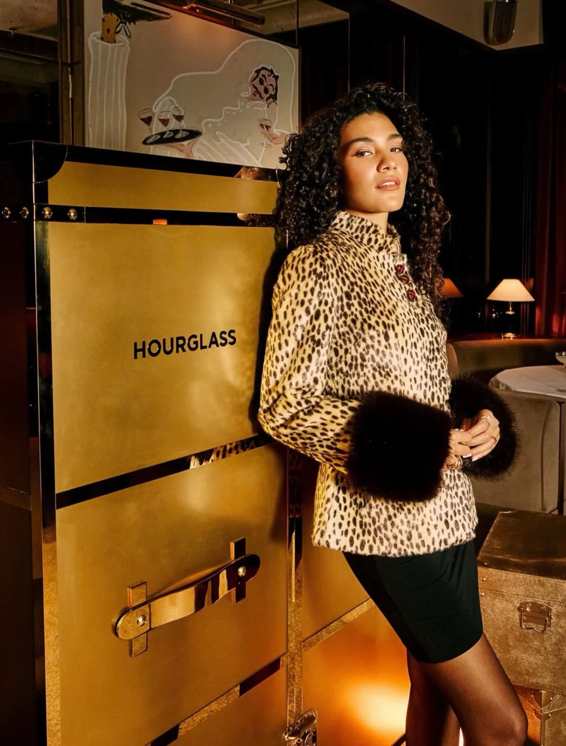 A person with curly hair wears a leopard print coat with dark fur cuffs and a black skirt, standing beside a large gold box labelled HOURGLASS in a stylish, warmly lit room that celebrates small fashion brands.