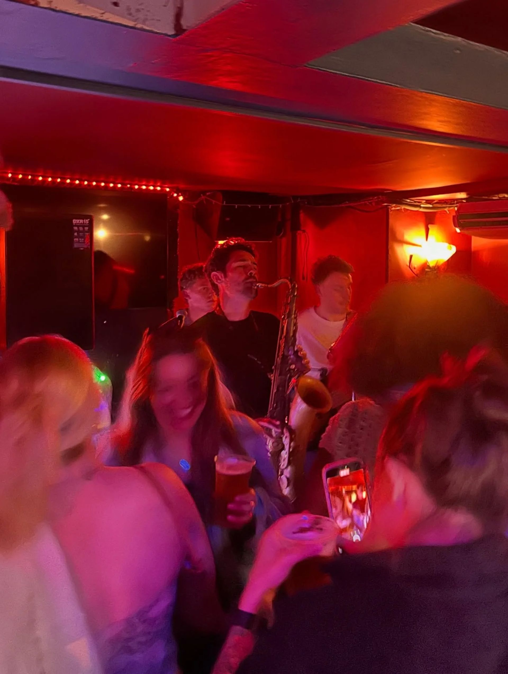 A lively bar scene with people dancing and socialising in dim lighting—a saxophonist performs as someone captures the moment on their mobile. Warm red lights illuminate one of the best things to do in Brighton.