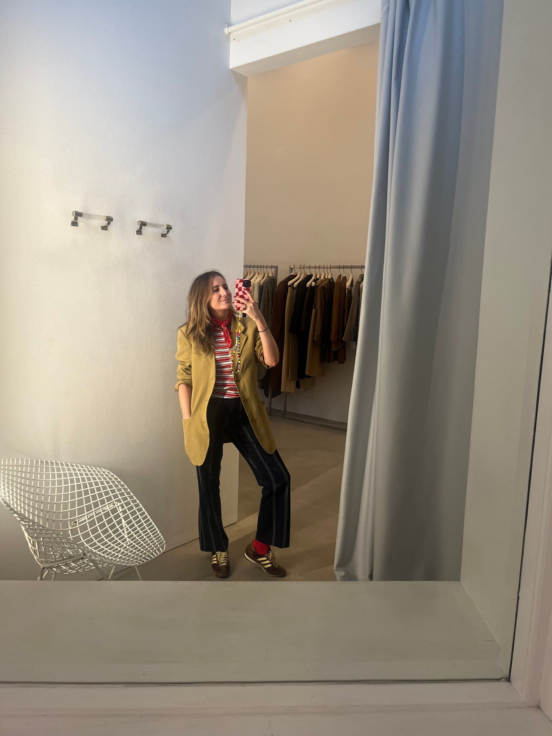 A woman takes a mirror selfie in a Copenhagen clothing shop, wearing a tan blazer, red-and-white striped shirt, dark trousers, and trainers. Behind her are hanging clothes and a white wire chair—perfect inspiration for 2026 travel looks.