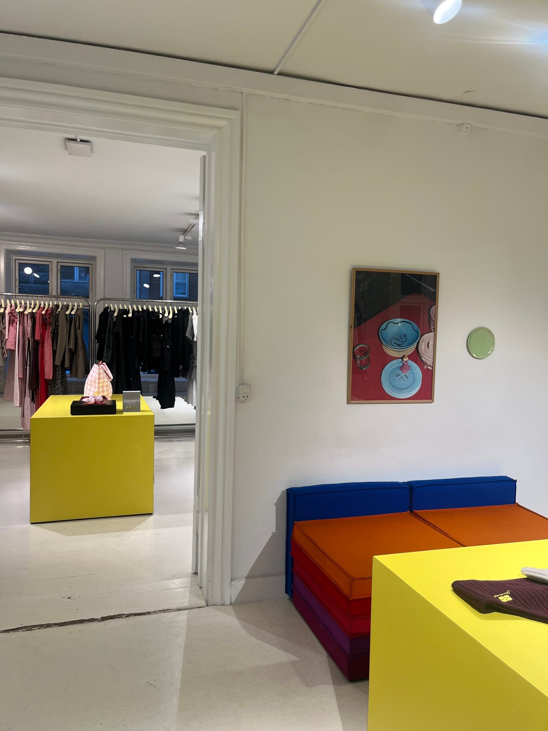 A modern clothes shop in Copenhagen with white walls, yellow display tables, and colourful bench seating—perfect for travel 2026 inspiration. A framed artwork and a small round mirror decorate the space’s contemporary charm.