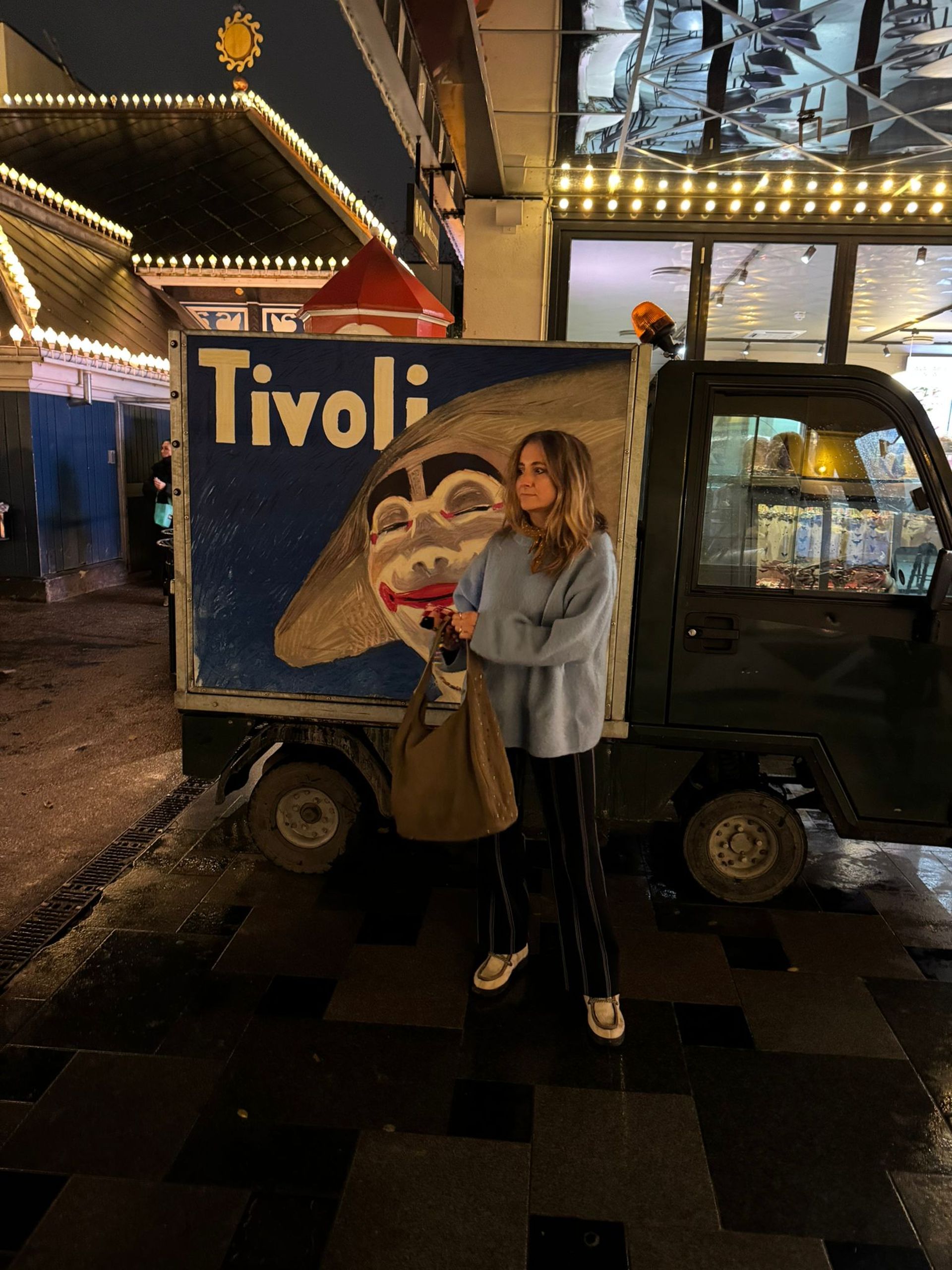 A woman stands at night in front of a small lorry with a large Tivoli sign and a painted clown face in Copenhagen. She wears a light blue jumper, holds a brown bag, and the area is lit by fairy lights—perfect for your 2026 travel memories.