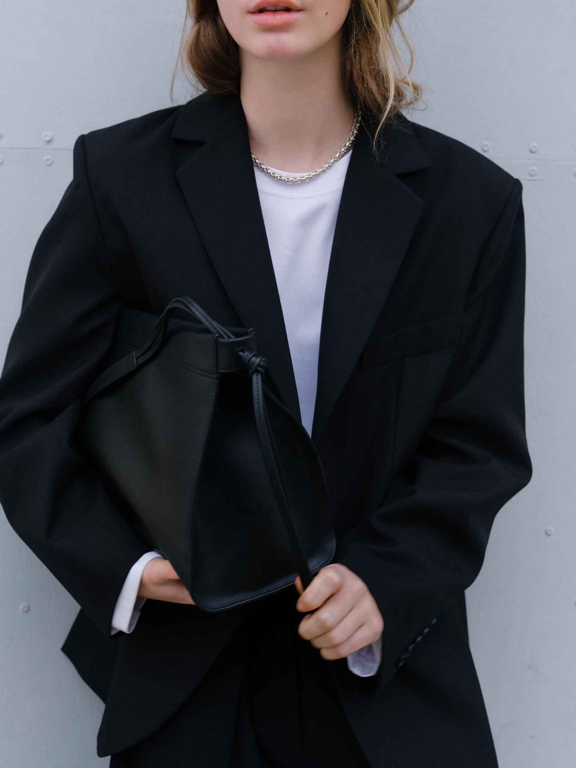 A person, styled like a fashion editor, wears a black high street blazer over a white shirt and holds a large black handbag. Only the lower part of their face is visible as they stand against a light grey background.