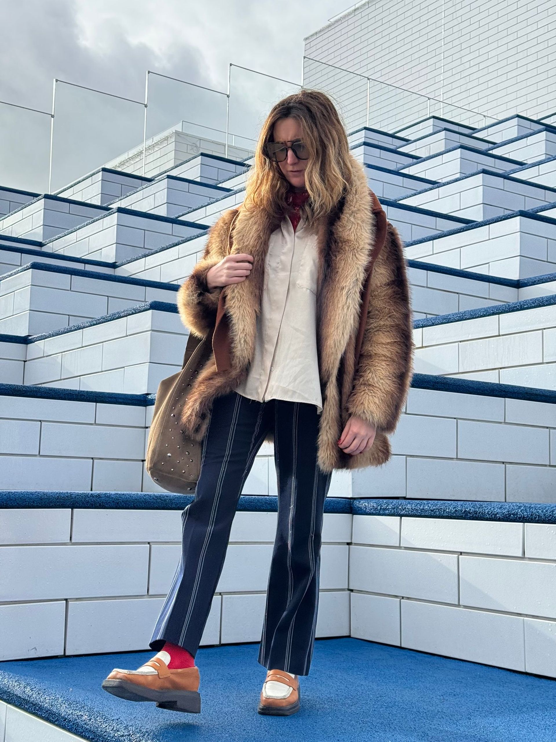 Showcasing Life in Fashion, a person wearing a faux fur coat, white shirt, striped trousers, and loafers stands on modern white and blue tiled steps. Wavy hair, sunglasses, and a brown bag complete this Fashion Editor-inspired look.
