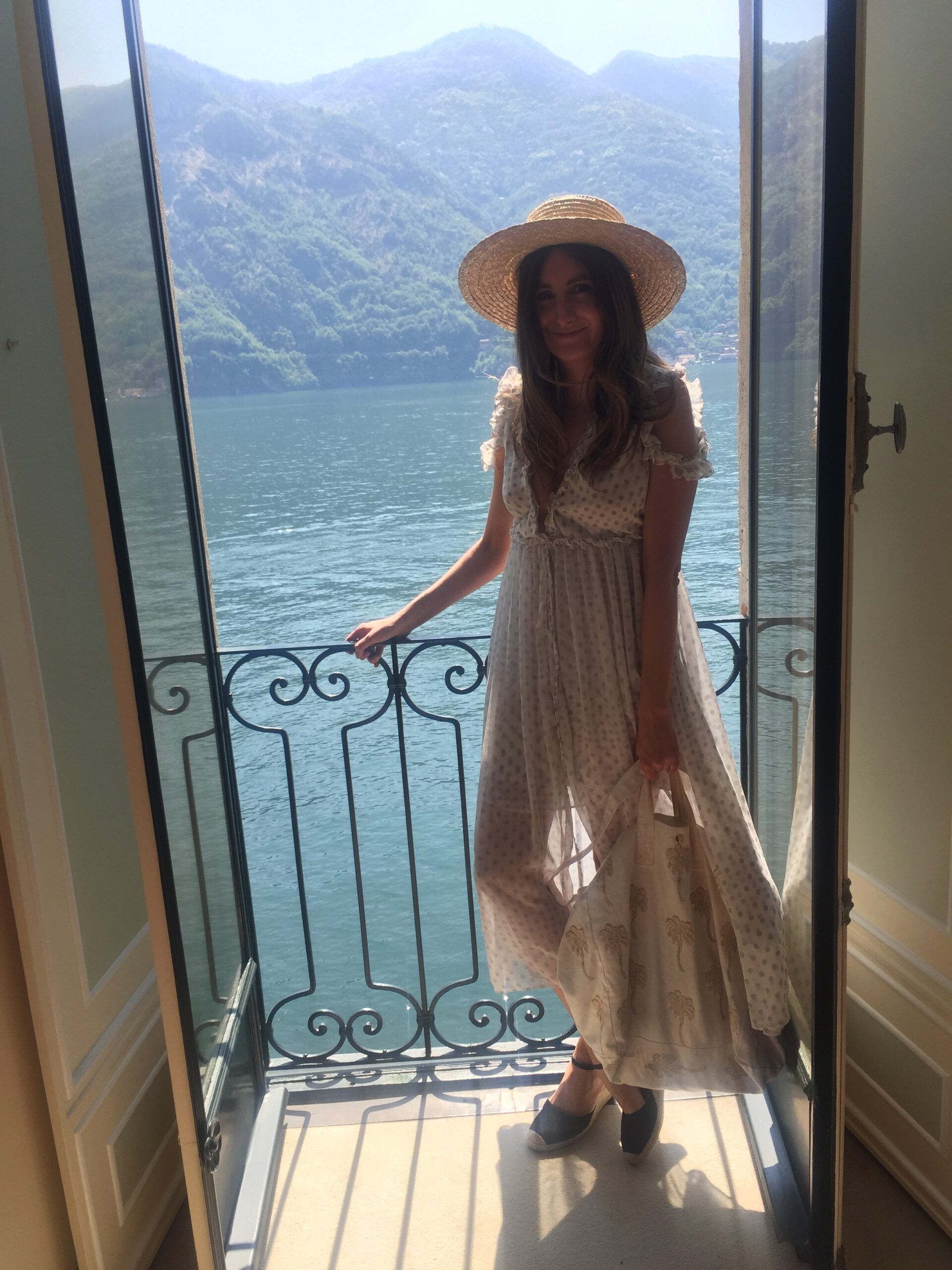 A woman, embodying Life in Fashion, stands smiling on a balcony in a light, floaty dress and straw hat, overlooking a tranquil lake and mountains, all bathed in soft natural sunlight.