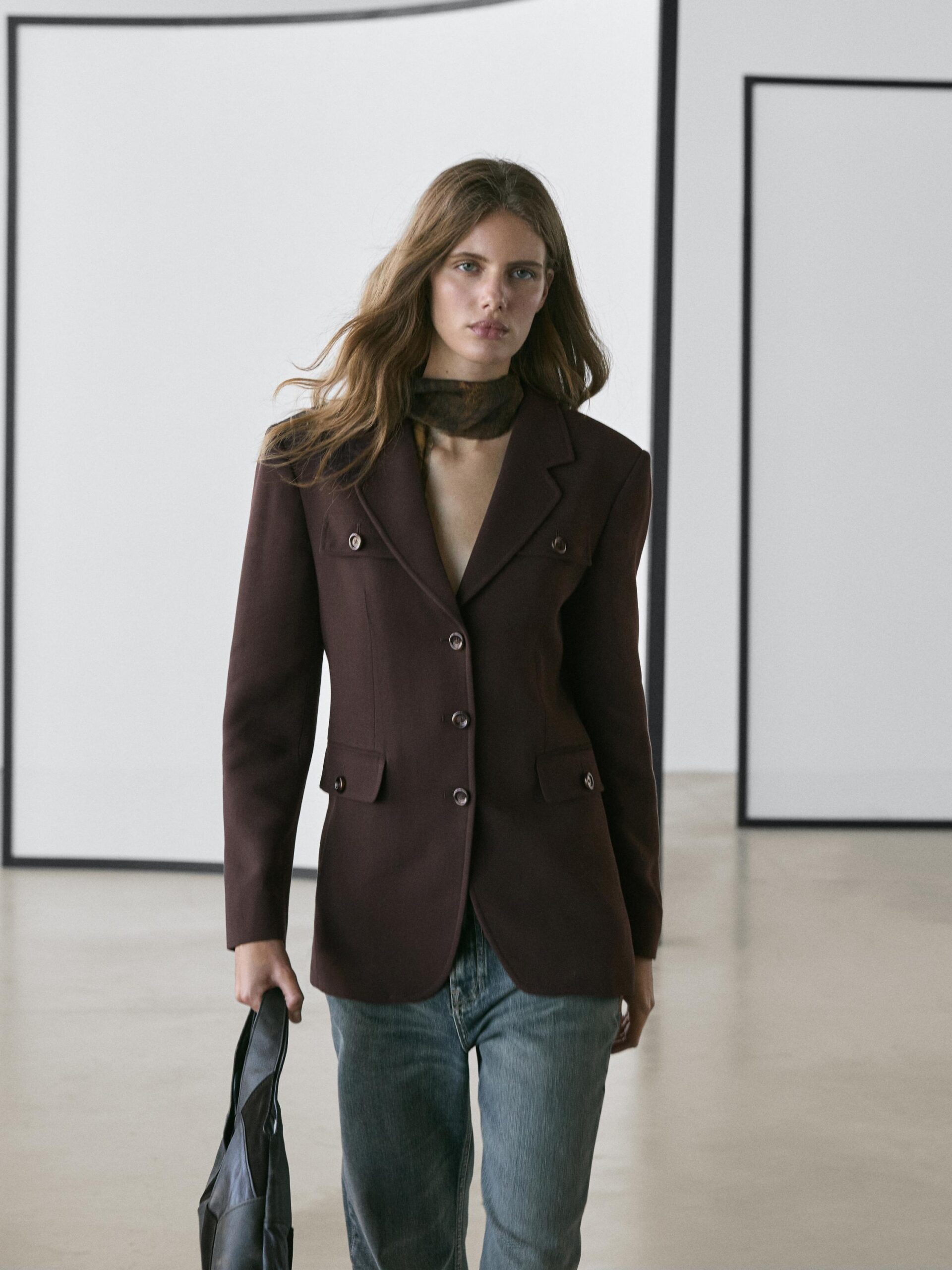 A woman with long hair, styled like a true Fashion Editor, wears a dark maroon high street blazer, a sheer scarf, and blue jeans. She holds a black bag whilst walking in a minimalist room with white walls and black rectangular frames.