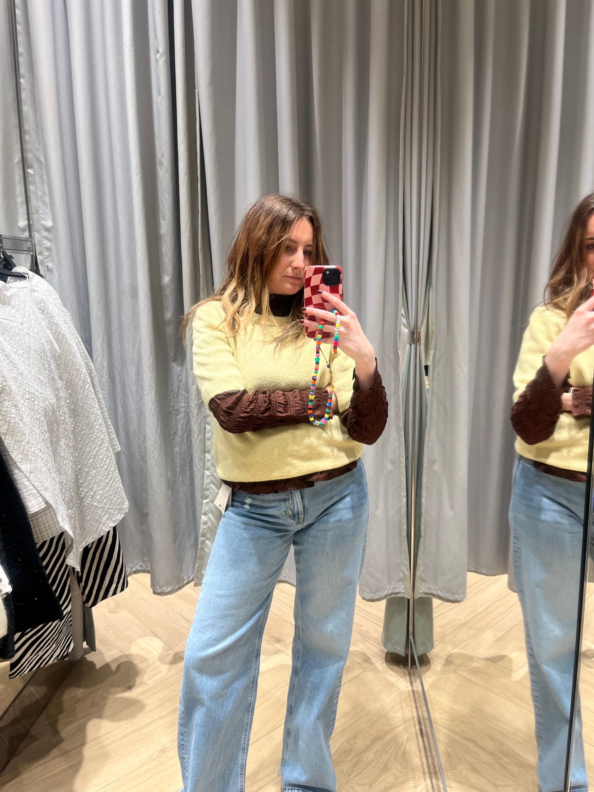 A woman poses for a mirror selfie in a fitting room, wearing an H&M new season yellow jumper with brown sleeves and light blue jeans. Clothing rails and grey curtains are visible in the background.