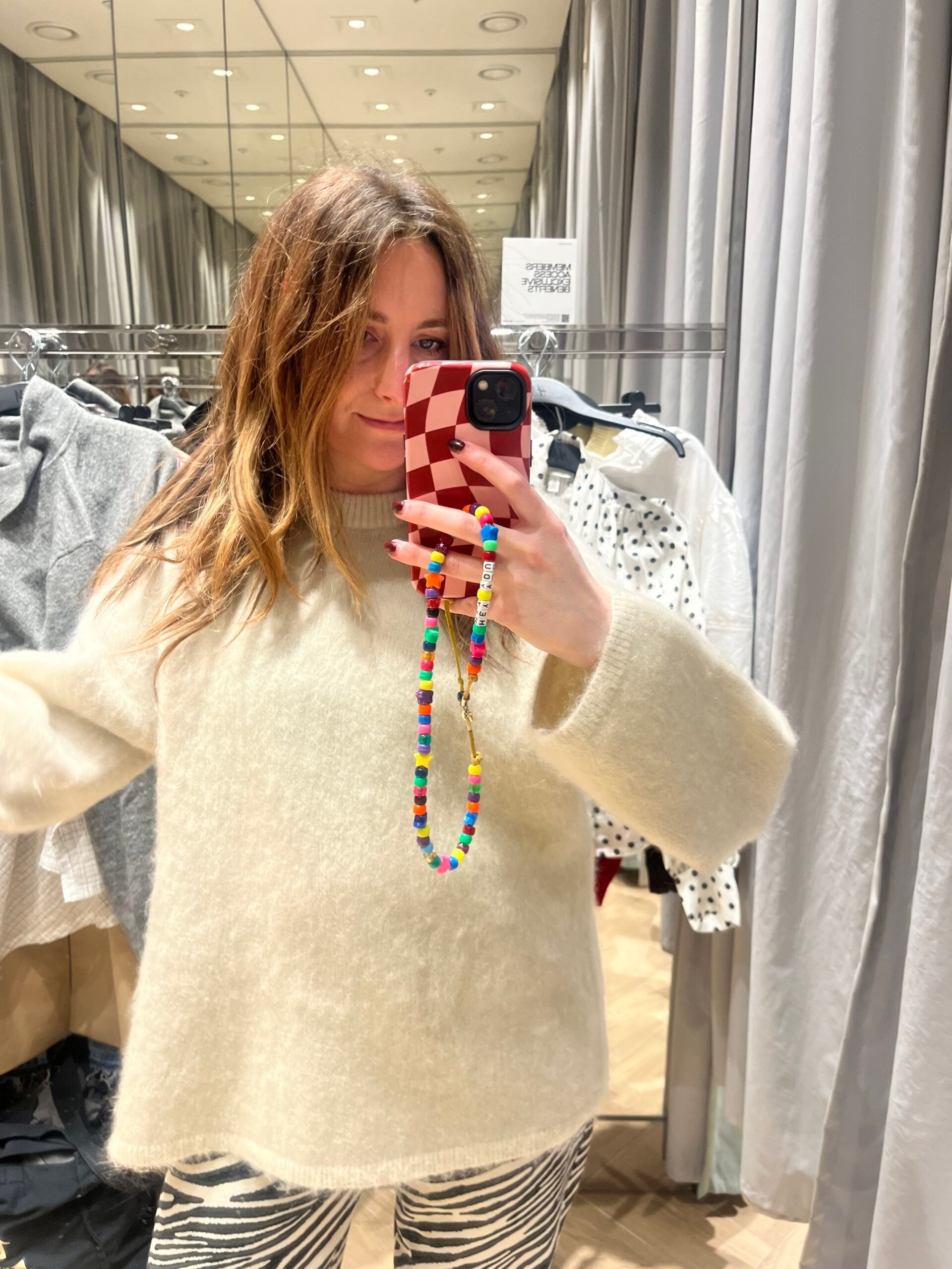 A woman with long hair takes a mirror selfie in a changing room, wearing H&M new season zebra-print trousers and a cream jumper. Her red and white chequered phone case has a colourful beaded strap, and clothes hang behind her.