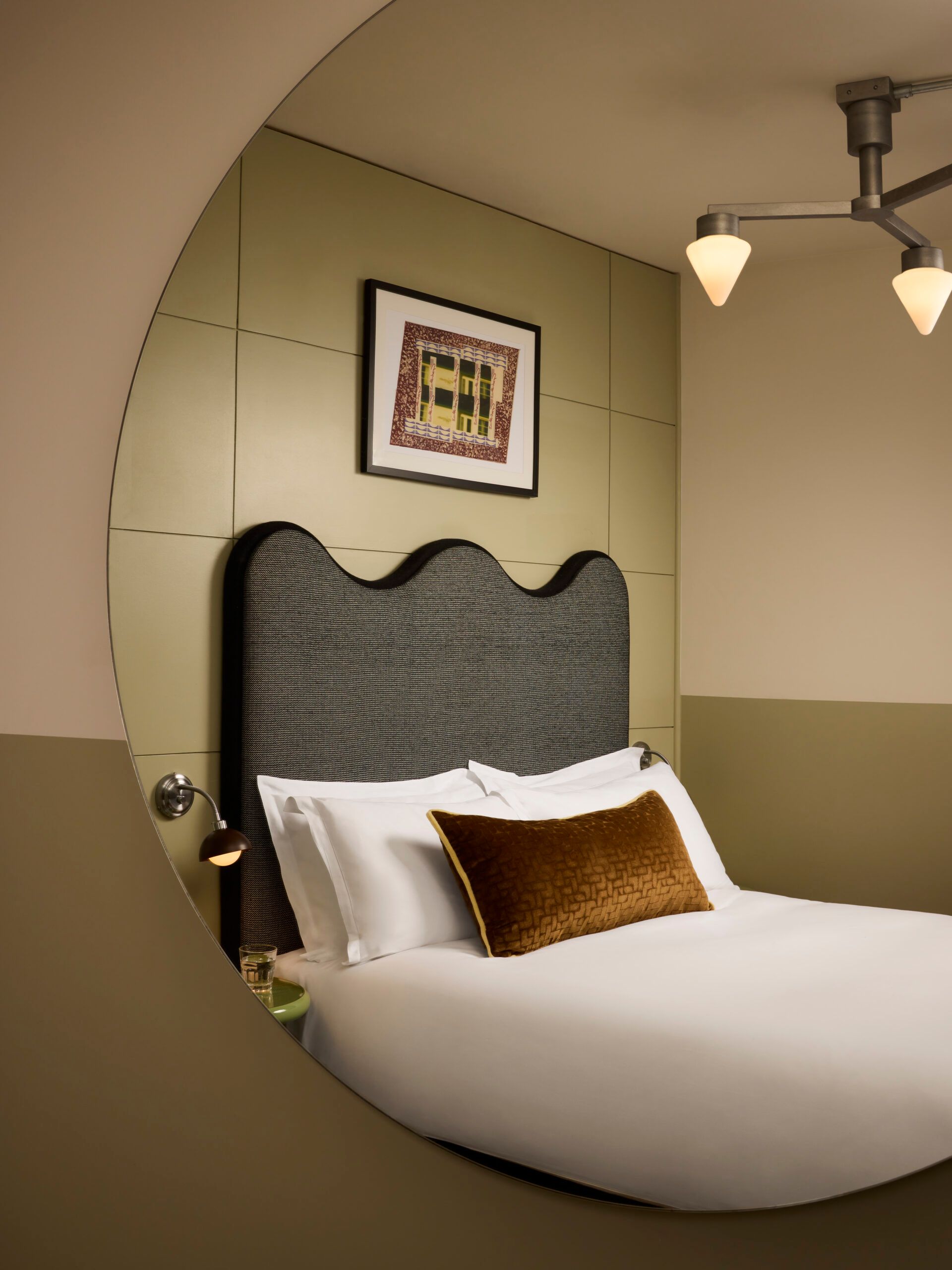 A modern hotel room at Hoxton Shoreditch in London is reflected in a round mirror, showcasing a bed with white linen, a gold-brown accent cushion, a wavy dark grey headboard, wall art, and a contemporary ceiling light fitting.