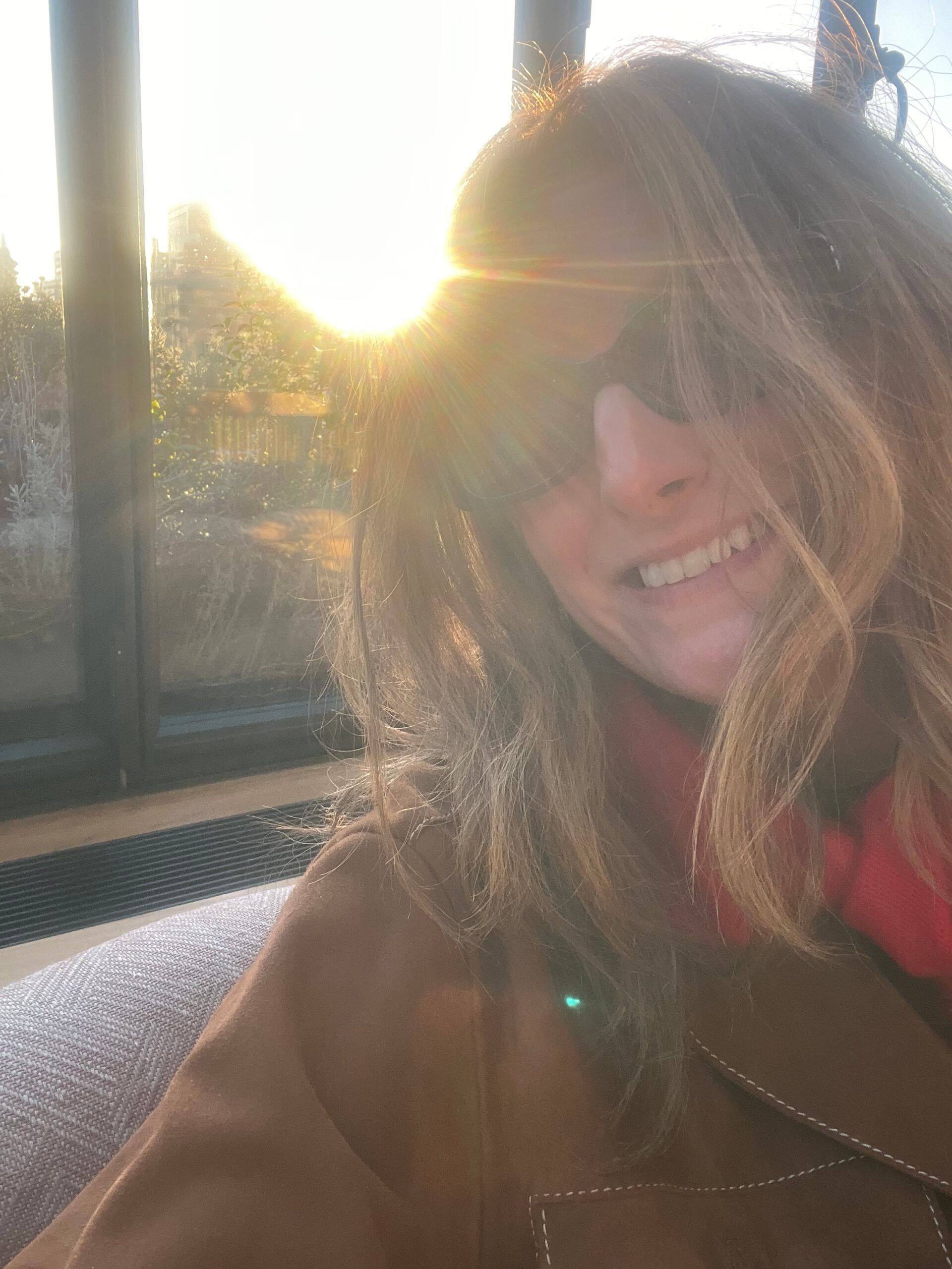 A woman wearing sunglasses and a brown coat smiles at the camera inside The Hoxton, with sunlight streaming through a window behind her, creating a warm and bright atmosphere.
