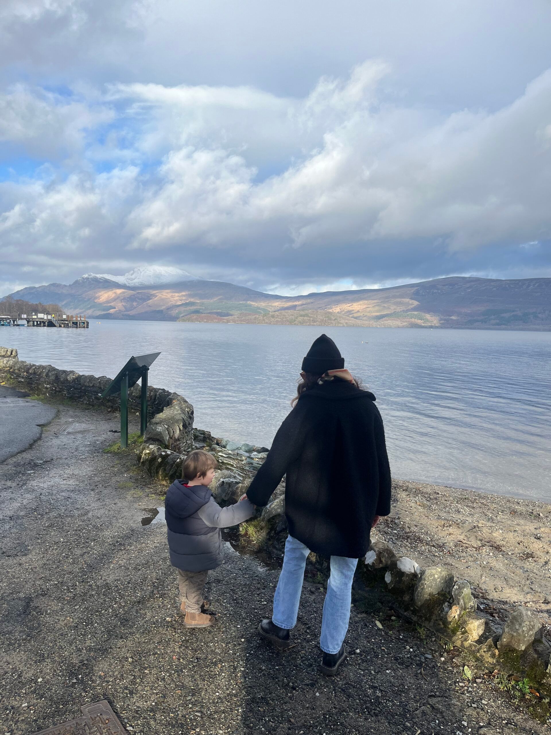 A woman and a young child, both in winter clothes, hold hands as they walk along a path by a calm lake—an intimate moment of motherhood set against mountains and clouds in the background.