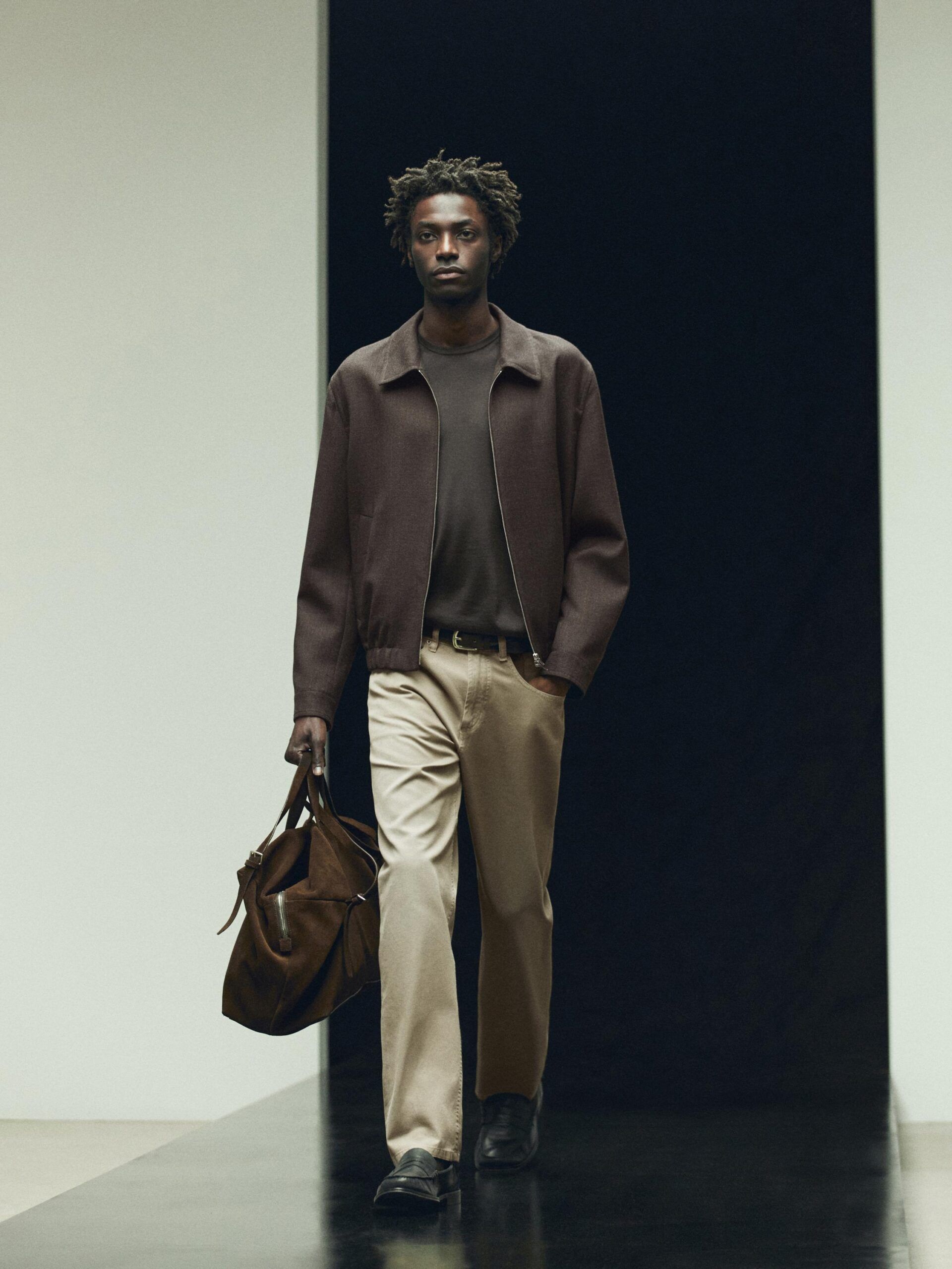 A man with short curly hair walks on a catwalk wearing a stylish brown men's jacket, dark shirt, beige trousers, and black shoes, carrying a large brown bag in his left hand. The background is split between white and black.