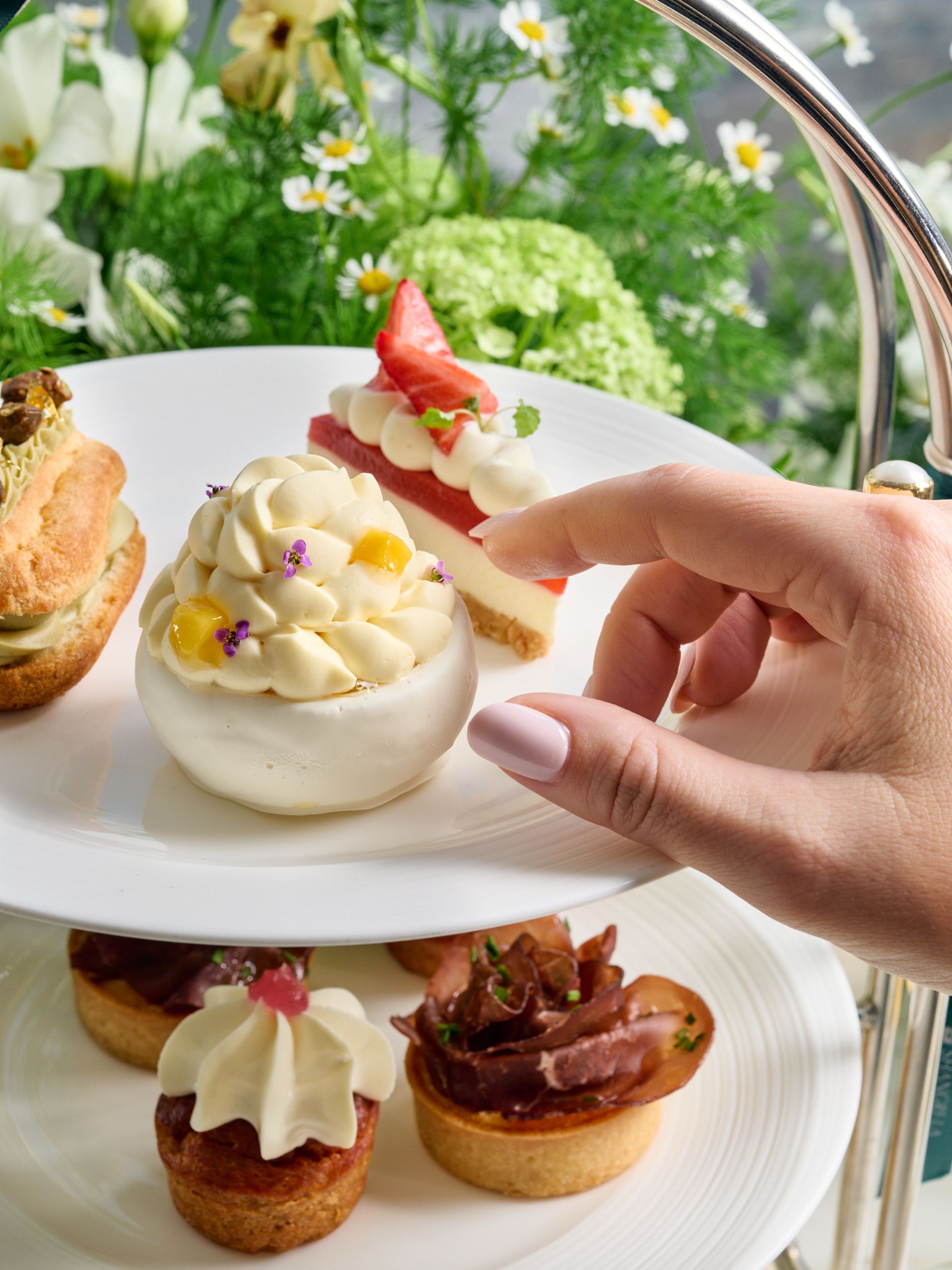 A hand reaches for a delicate white pudding topped with cream and fruit on a tiered tray, surrounded by assorted pastries and flowers—capturing the charm of March pop-ups.