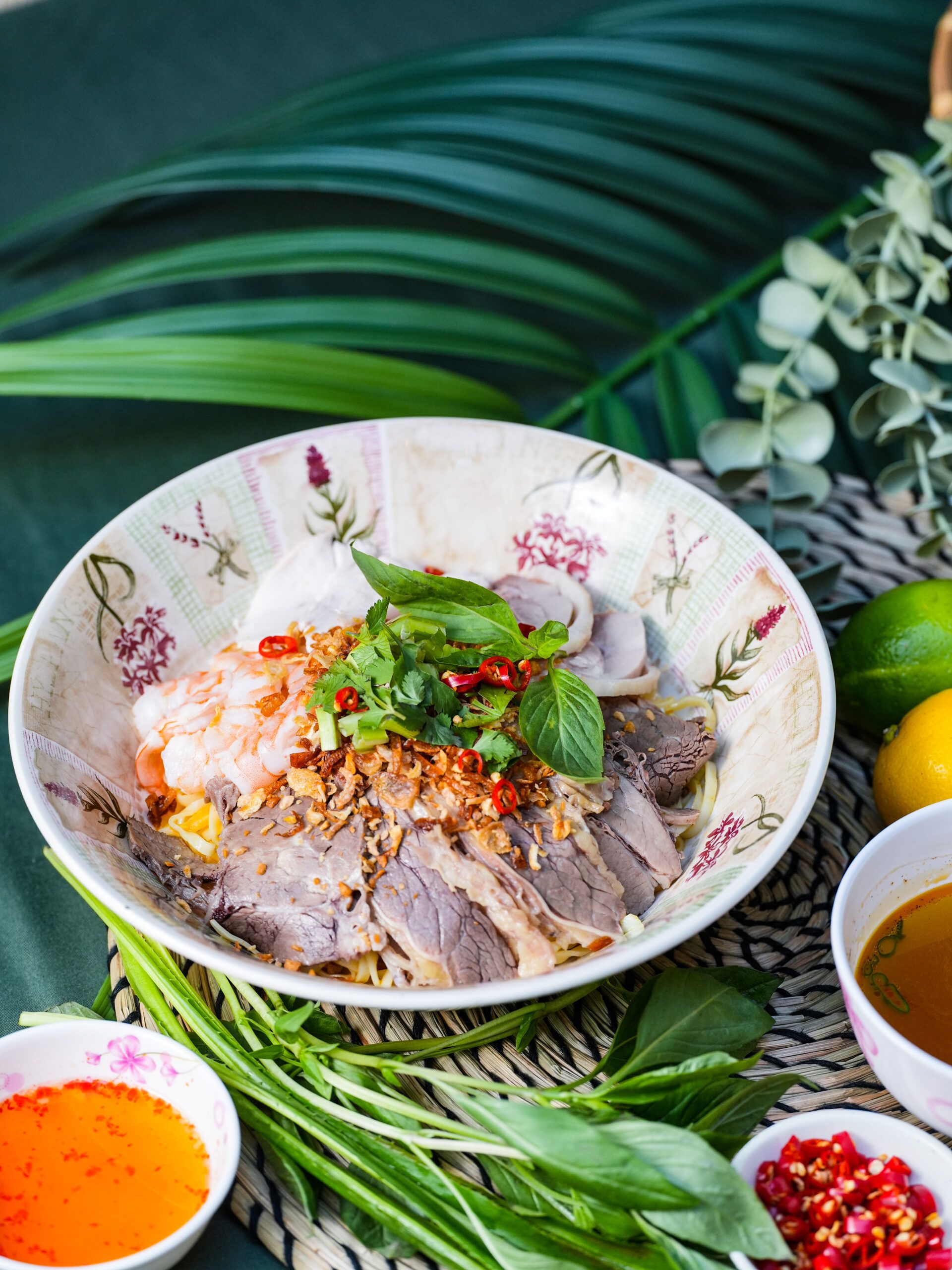 A bowl of Vietnamese noodle salad topped with sliced beef, prawns, herbs, chopped chilli, and fried shallots, surrounded by fresh greens, lime, dipping sauces, and leafy table decor in a style reminiscent of Hannah Crosbie.