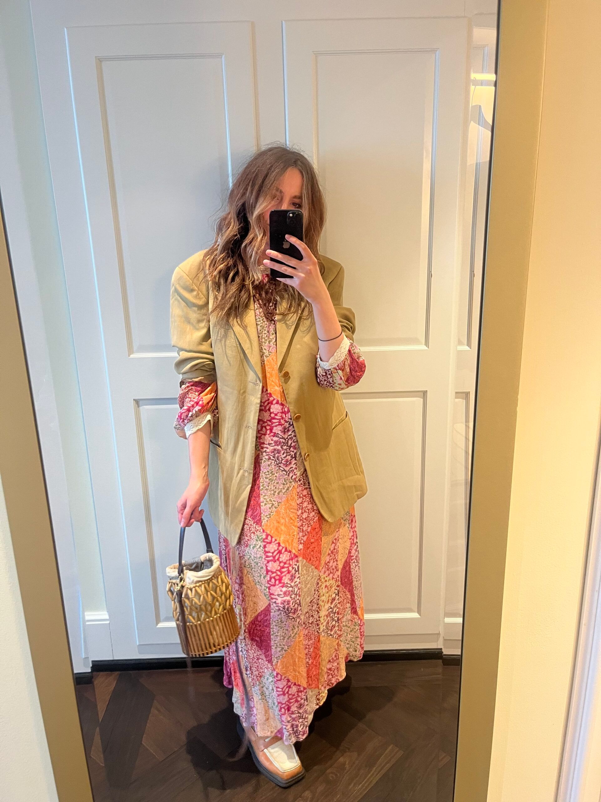 A person takes a mirror selfie at the Dolder Grand in Zurich, wearing a patchwork floral maxi dress, beige blazer, and metallic shoes. They hold a woven handbag; wavy hair frames their face against a white panelled door in this fairy-tale hotel setting.