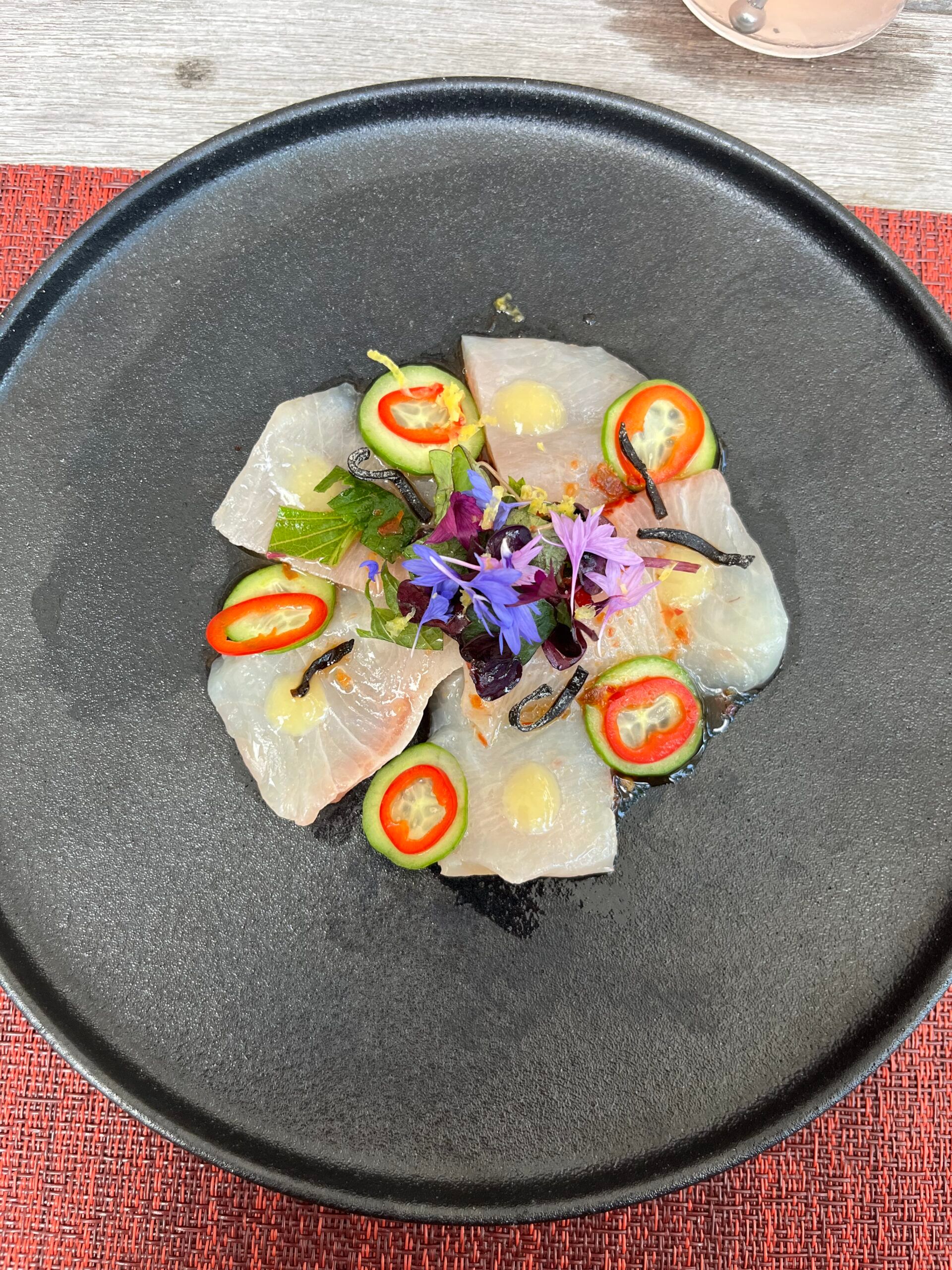 Thin slices of raw white fish arranged on a black plate, garnished with red chilli slices, edible flowers, herbs, and light sauce—an elegant dish reminiscent of fine dining at Zurich’s iconic Dolder Grand fairytale hotel.