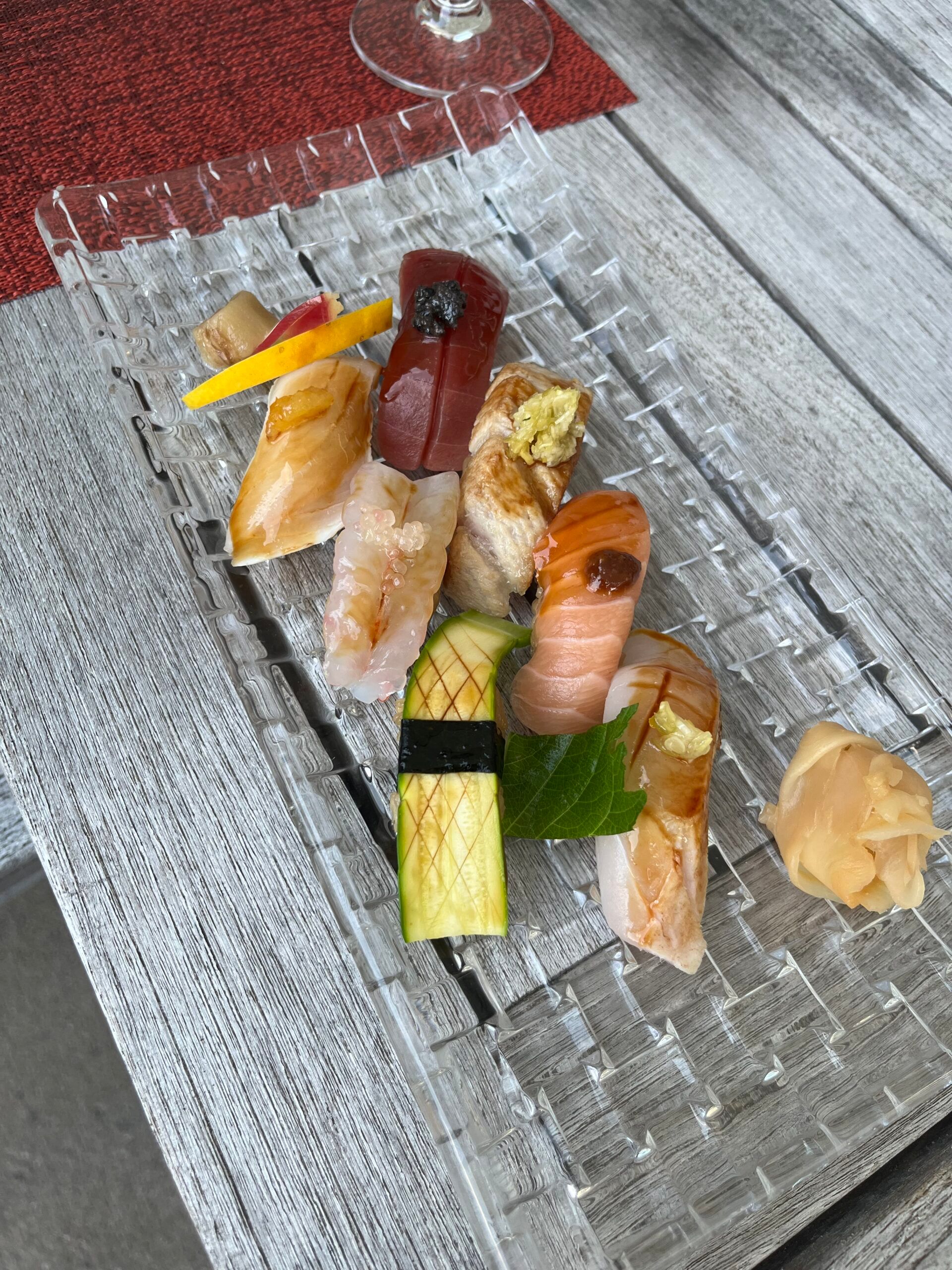 A clear rectangular plate with assorted sushi pieces, including nigiri topped with fish, prawns, and vegetables, garnished with ginger, sits on a wooden table at the Dolder Grand in Zurich—a fairytale hotel—with a red placemat and part of a wine glass visible.