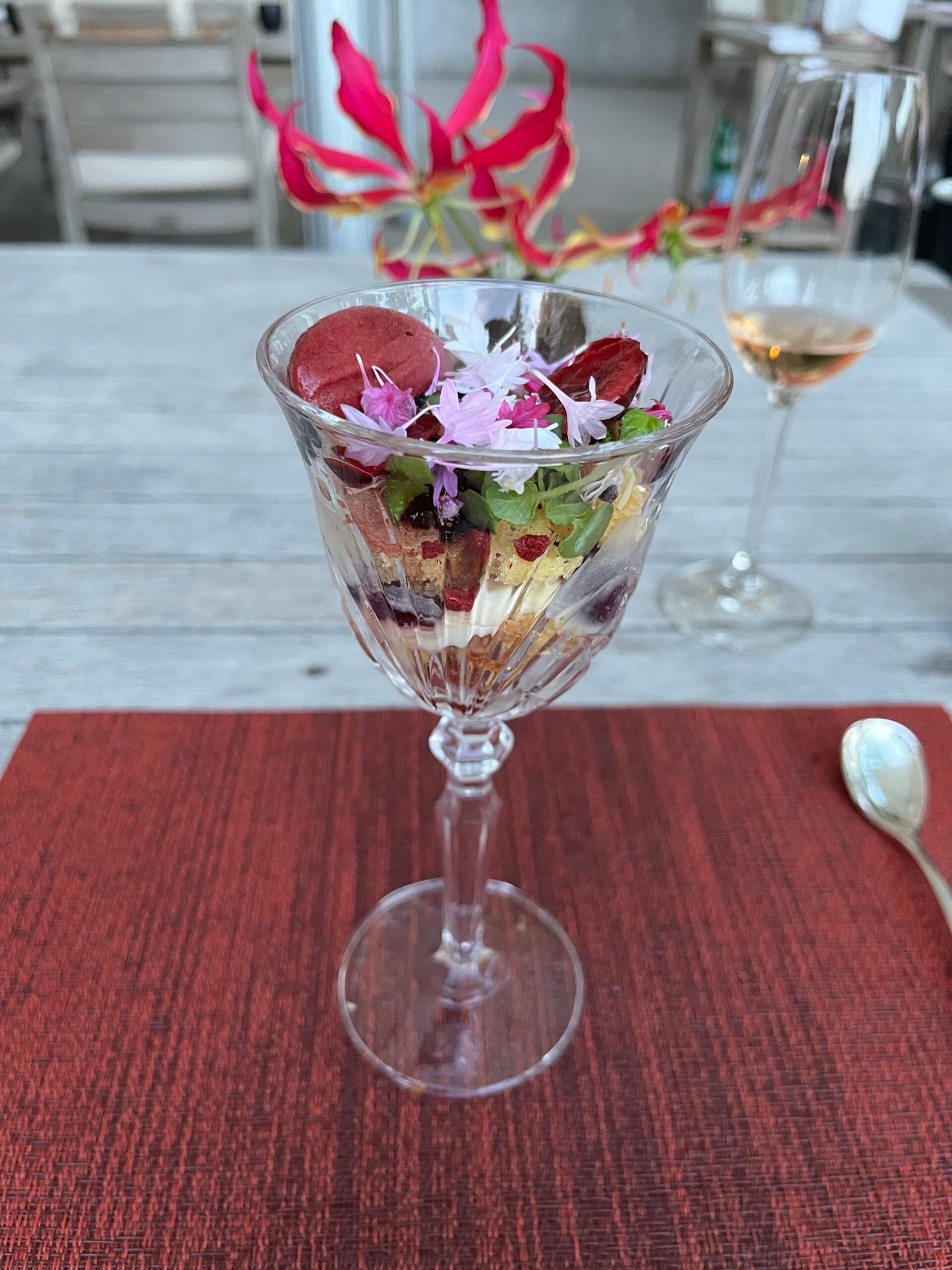 A crystal glass filled with a layered pudding topped with edible flowers and herbs sits on a red placemat at the Fairytale Hotel Dolder Grand. A wine glass and a pink flower in a vase are in the background on a wooden table in Zurich.