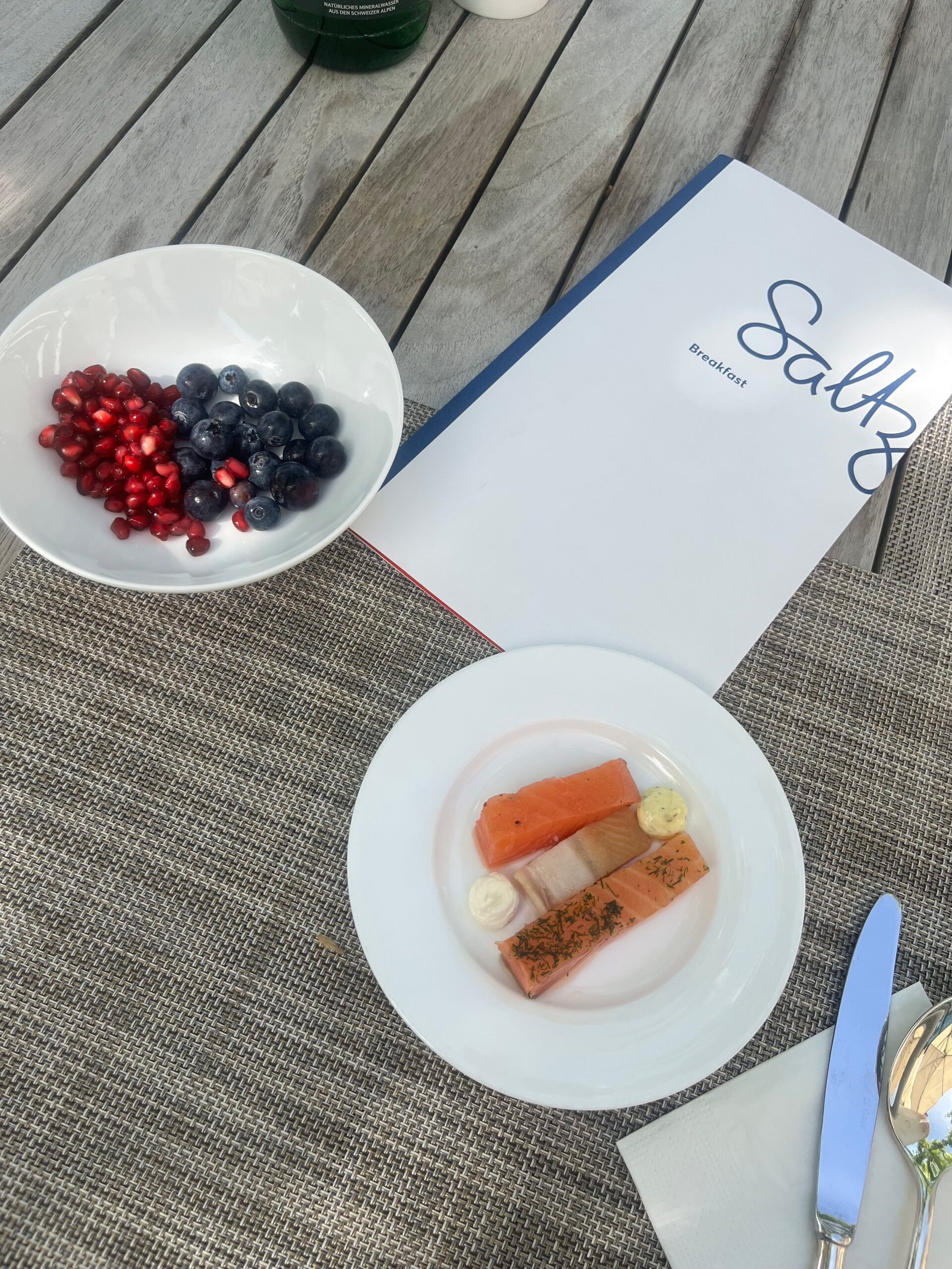 A breakfast setting at the Dolder Grand in Zurich, featuring a bowl of pomegranate seeds and blueberries, smoked salmon with garnishes, cutlery, and a menu labelled “Saltz” on a wooden table—a scene from a fairytale hotel.