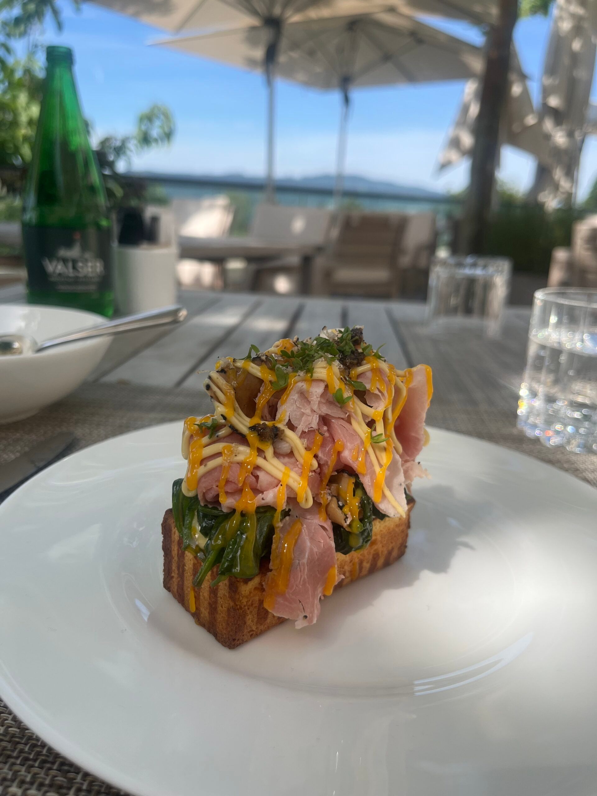An open sandwich topped with ham, grated cheese, and salad leaves sits on a white plate at an outdoor restaurant table at the fairytale hotel Dolder Grand in Zurich, with a water bottle, glass, and patio umbrellas in the background.